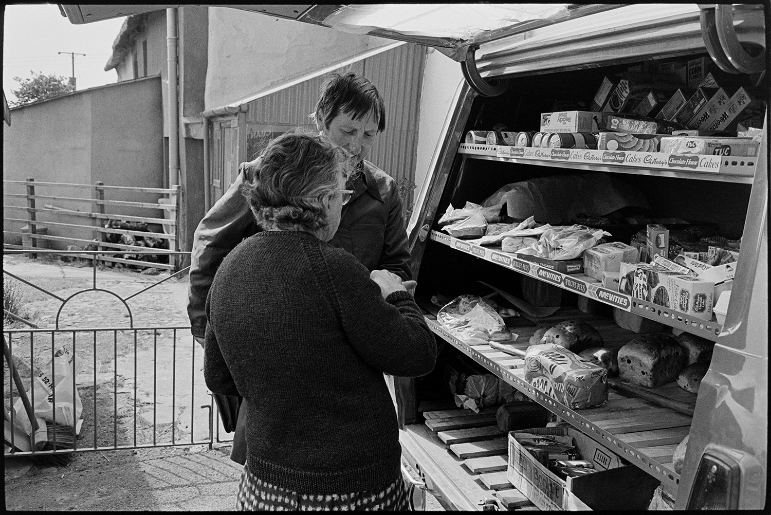 Women buying food from mobile grocers van. <br />
[Miss Audrey Bowden buying items from a Grocer's van in a street in Kings Nympton. She is paying a man. Items on the van include bread, biscuits, cans and scones.]