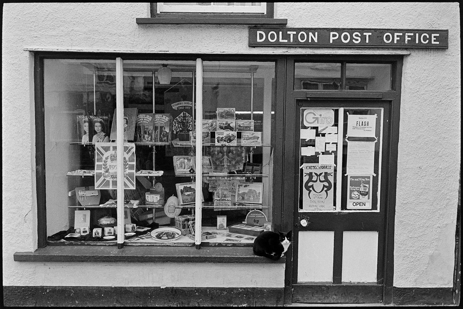 Front of Post Office with display of jubilee souvenirs. 
[The front window of Dolton Post Office decorated with royal souvenirs including plates and tea towels for Queen Elizabeth II Silver Jubilee. A cat is sat on the window sill.]
