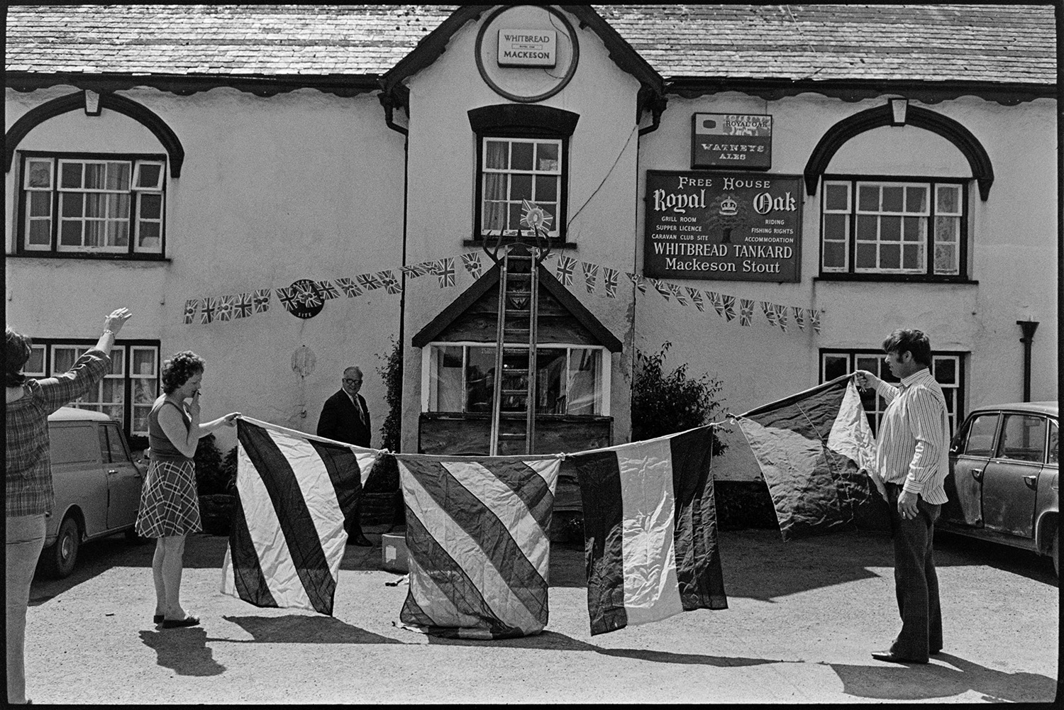 Putting up flags for jubilee on village pub. 
[Men and women decorating the Royal Oak pub in Dolton with flags, including Union Jack flags for Queen Elizabeth II Silver Jubilee. A ladder is leaning against the pub in the background.]