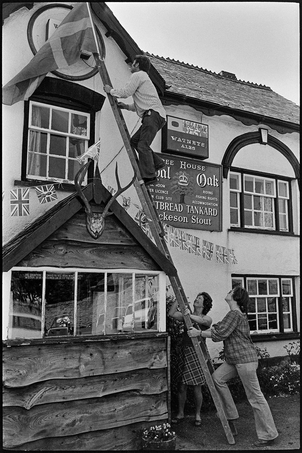 Putting up flags for jubilee on village pub. 
[A man up a ladder decorating the Royal Oak pub in Dolton with flags for Queen Elizabeth II Silver Jubilee. A man and a woman are holding the bottom of the ladder. The pub porch has a pair of antlers mounted on it.]