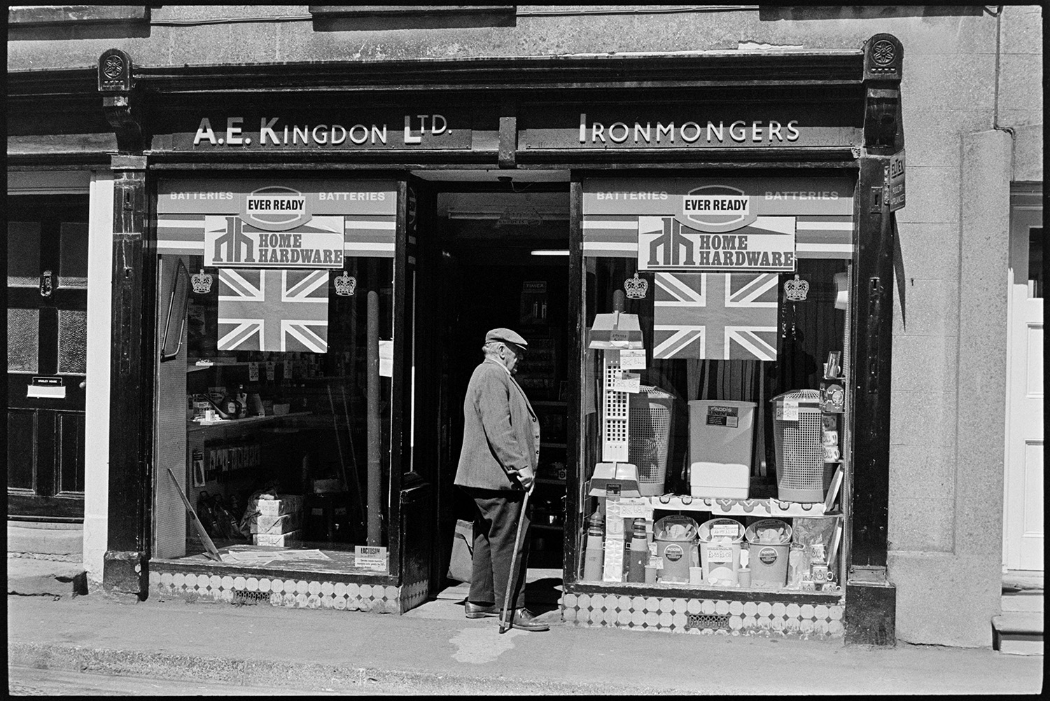 Ironmongers shop decorated for jubilee. 
[A man stood outside A E Kingdon, Ironmongers shop in Fore Street Chulmleigh. He is looking at the front window display which is decorated with Union Jack flags for Queen Elizabeth II Silver Jubilee.]