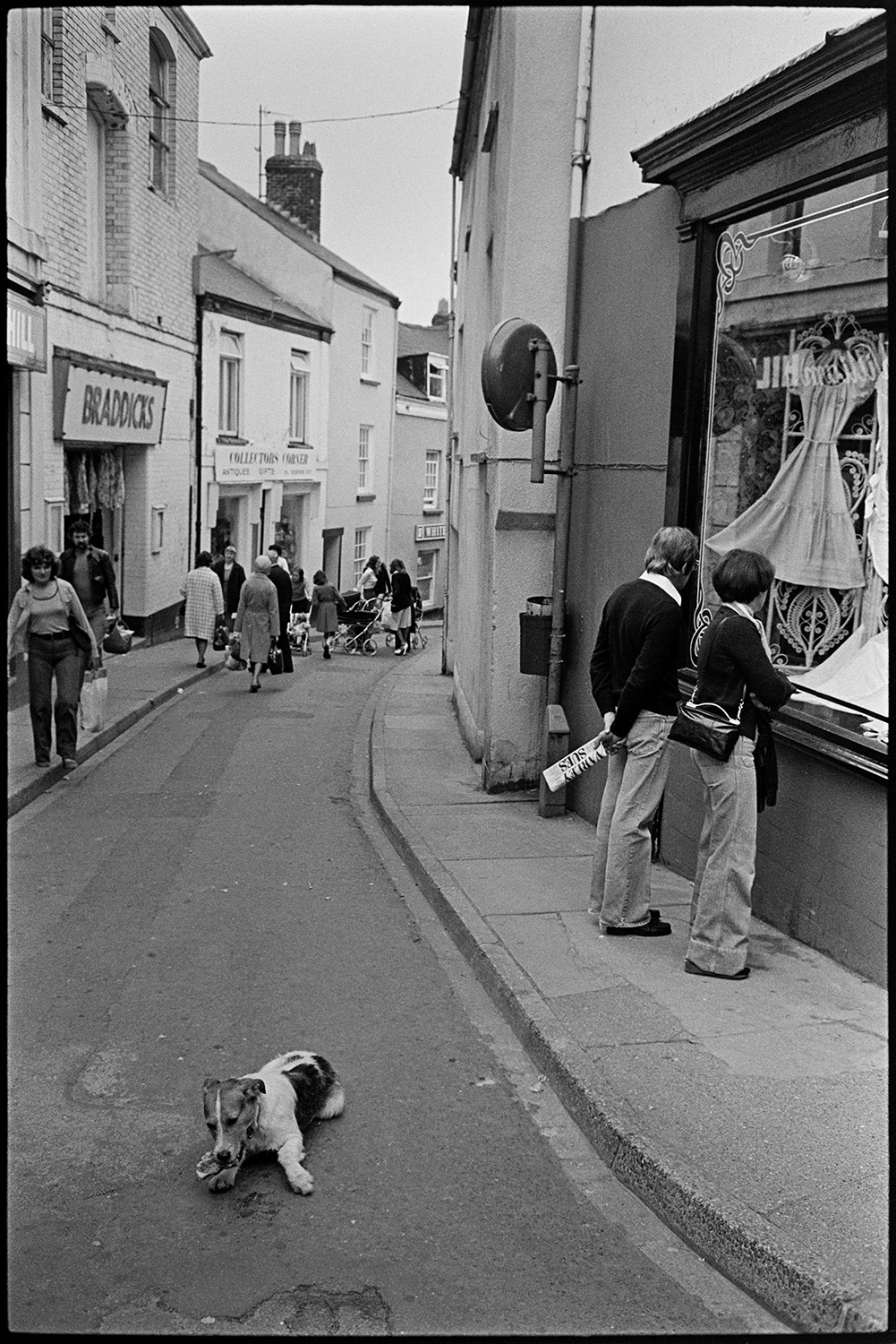 Street scenes with shop burnt out, dog eating bone !! 
[Shoppers in Bideford High Street looking in shop windows. Shop fronts, including Braddicks can be seen. In the foreground a dog is eating a bone in the middle of the street.]