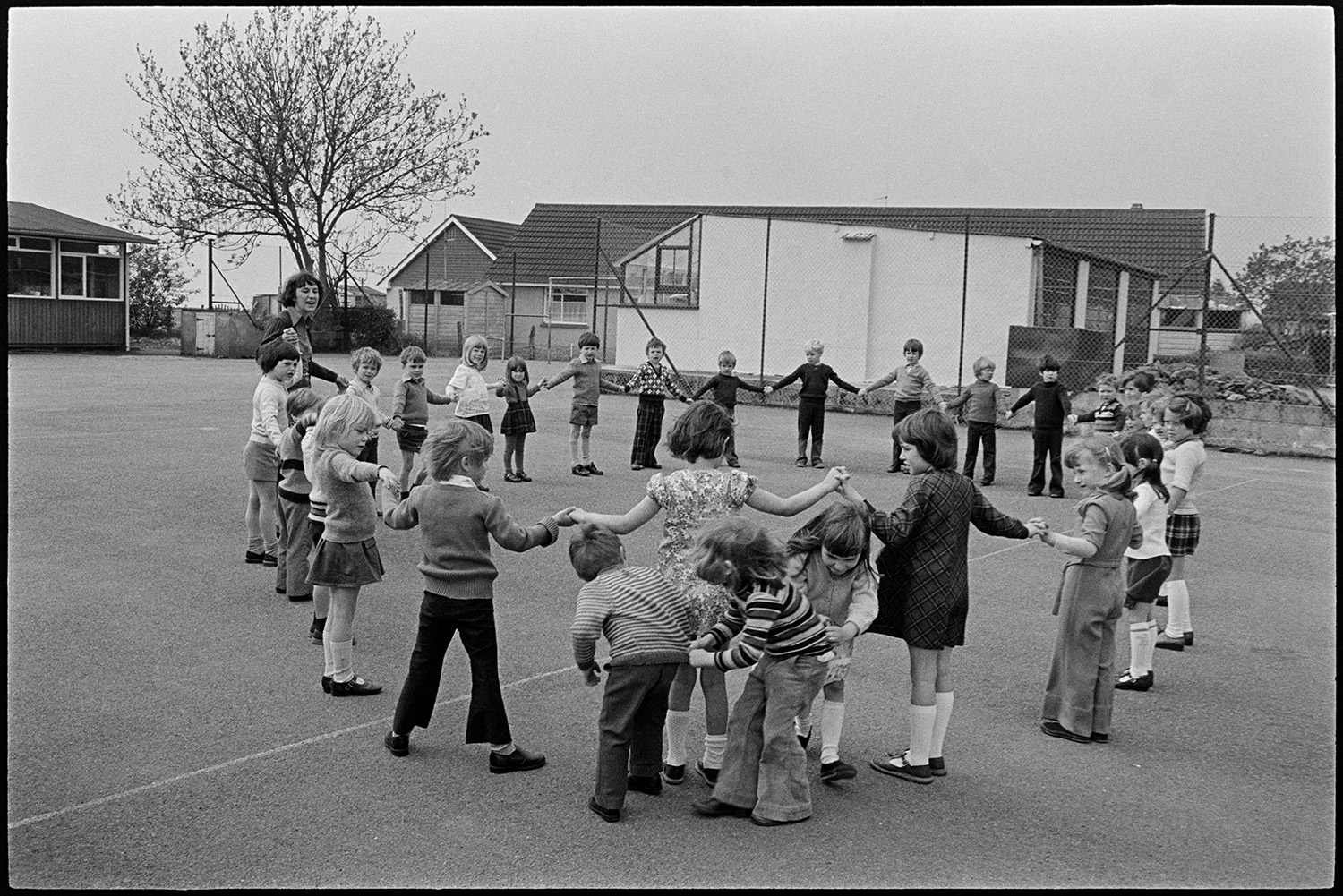 Children playing in school playground. 
[School children and a teacher playing in Beaford Primary School playground. They are holding hands in a circle while three other children weave in and out of the circle.]
