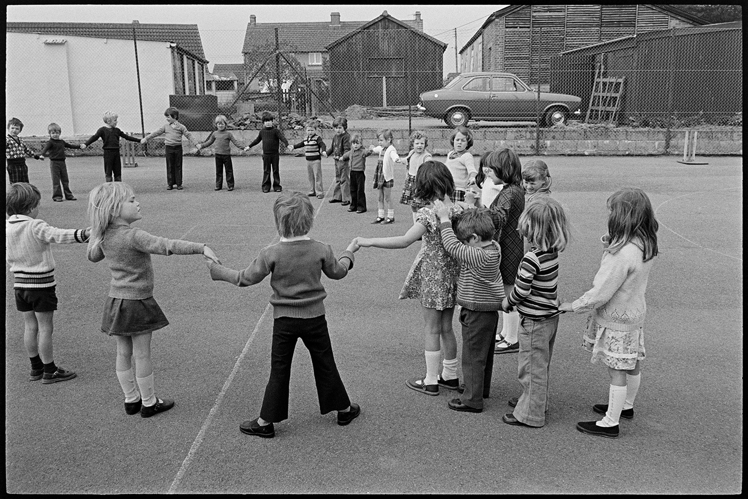 Children playing in school playground. 
[School children playing in Beaford Primary School playground. They are holding hands in a circle while other children line up behind the children in the circle.]