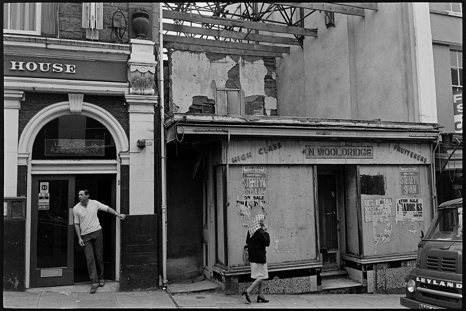 Street scenes with shop burnt out, dog eating bone !! 
[The burnt out shop front of N Wooldridge fruit shop in Bideford High Street. A woman is walking past and a man is coming out of the restaurant next door.]