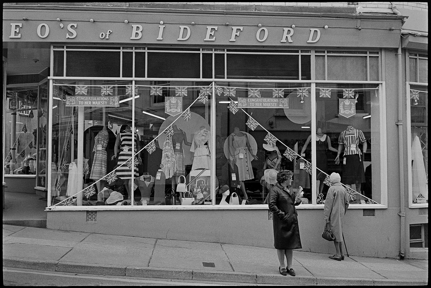 Clothes shop windows decorated for Jubilee. 
[The shop front window of Leo's of Bideford clothes shop decorated with Union Jack flags for Queen Elizabeth II Silver Jubilee. A woman I looking in the shop window while another is waiting to cross the road.]
