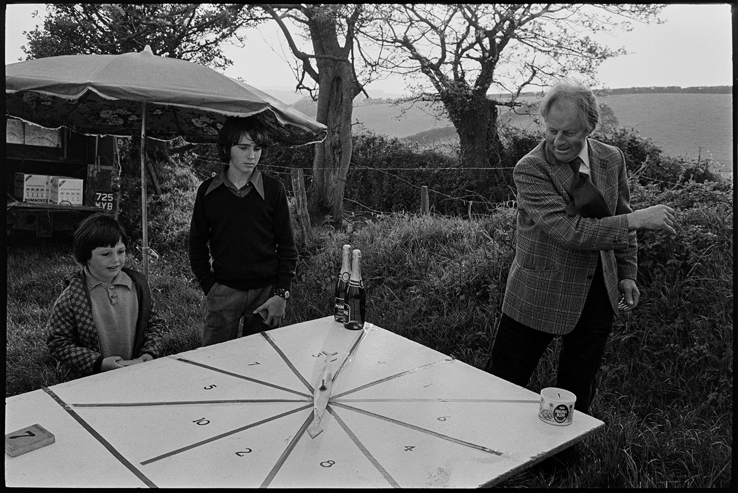 Clay pigeon shoot and games roulette for champagne. 
[Two boys stood under a parasol watching Alan Berry spin an arrow on a number wheel to try to win a bottle of champagne, at a clay pigeon shoot at Harepath, Beaford.]