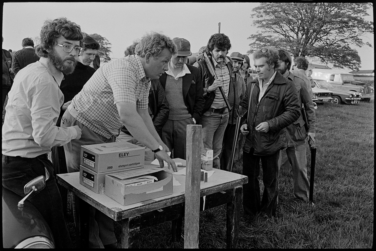 Clay pigeon shoot and games roulette for champagne. 
[Men collecting shotgun cartridges at a clay pigeon shoot at Harepath, Beaford. Parked cars can be seen in the background.]