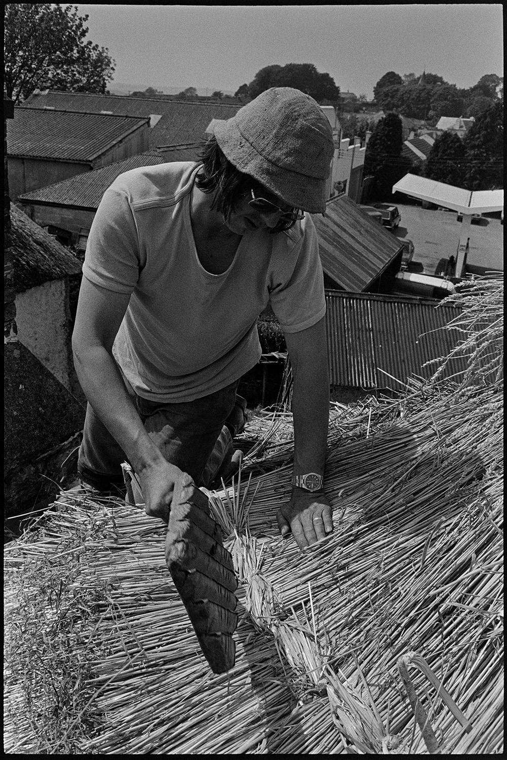 Thatcher thatching, taken from top of roof. 
[Nigel Gard thatching a roof in Beaford. He is using a wooden tool. The image is taken from the rooftop and other roofs can be seen in the background.]