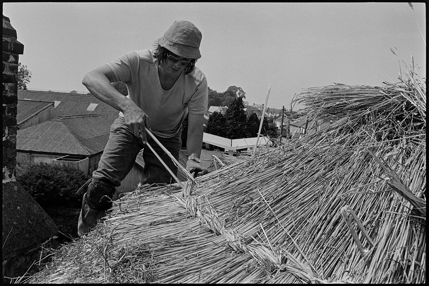 Thatcher thatching, taken from top of roof. 
[Nigel Ward thatching a roof in Beaford. He is using a spar to secure the thatch. The images is taken from the rooftop he is thatching and other roofs can be seen in the background.]