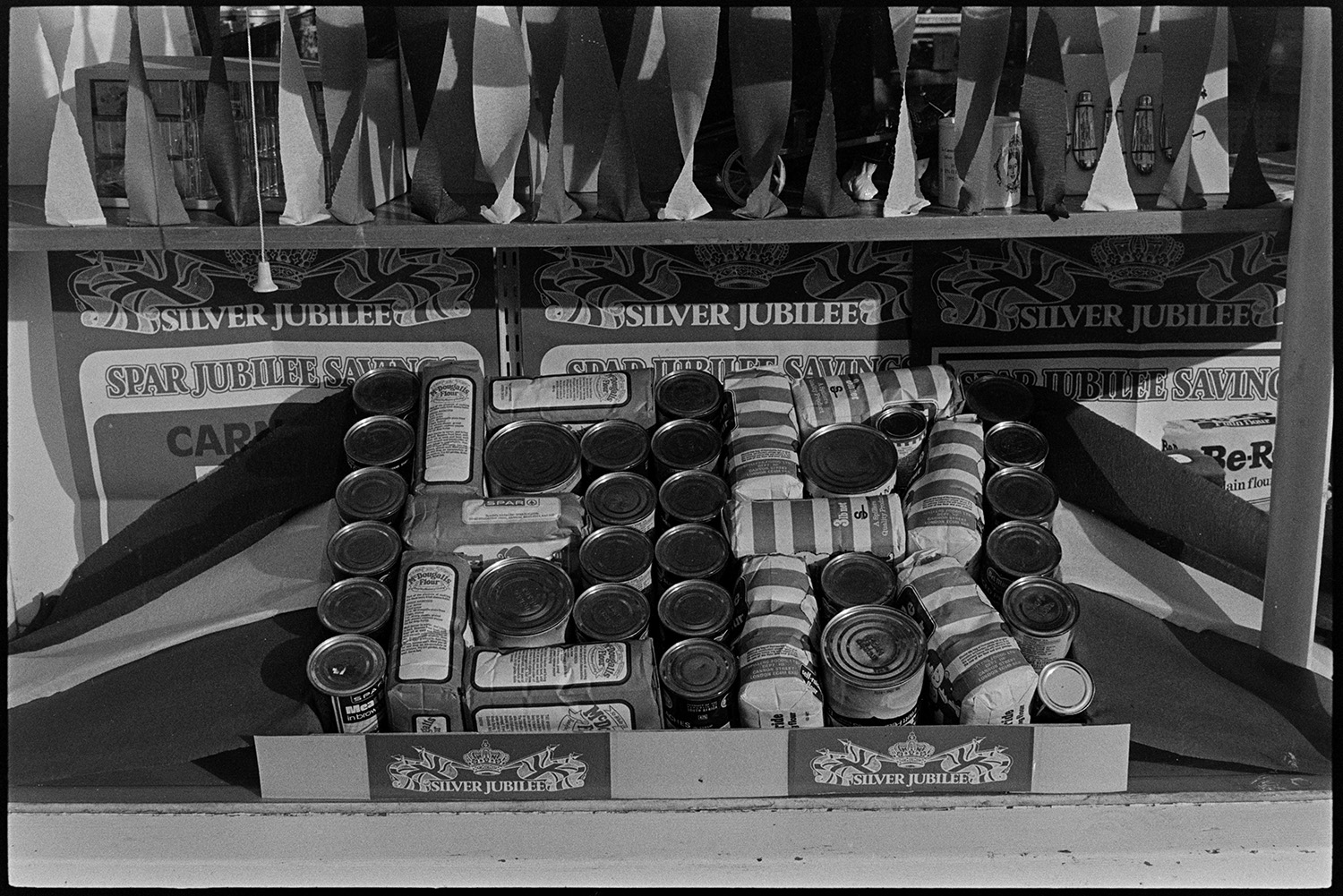 Village stores decorated with pattern of tins, for Jubilee. 
[A display of tins and bags of flour spelling 'E R' at London House Stores in Dolton, for Queen Elizabeth II Silver Jubilee.]