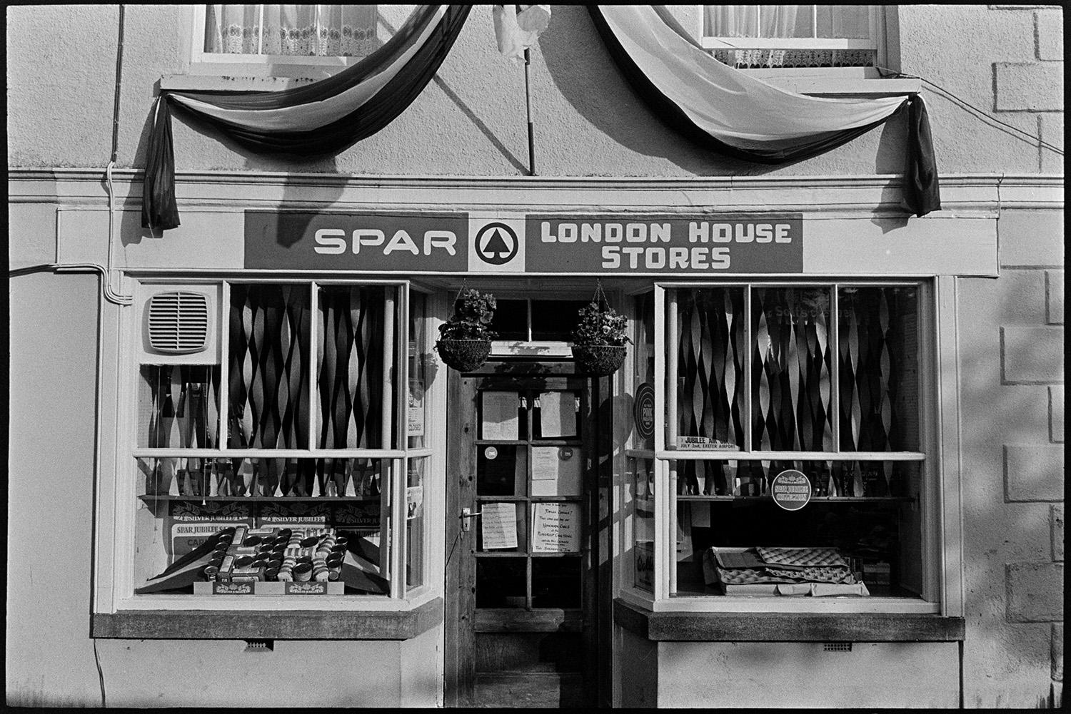 Village stores decorated with pattern of tins, for Jubilee. 
[The shop front window of London House Stores or Spar in Dolton. It is decorated for Queen Elizabeth II Silver Jubilee with the letters 'E R' spelt out in a display of tins and bags of flour. Hanging baskets are also above the shop door.]