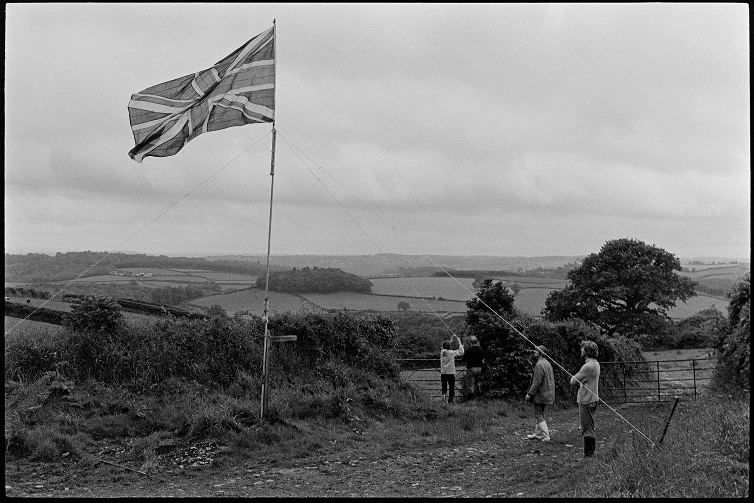 Hoisting the Jubilee flag in the countryside. 
[Three members of the Berry family hoisting a Union Jack flag by a field at South Harepath, Beaford, for Queen Elizabeth II Silver Jubilee.]