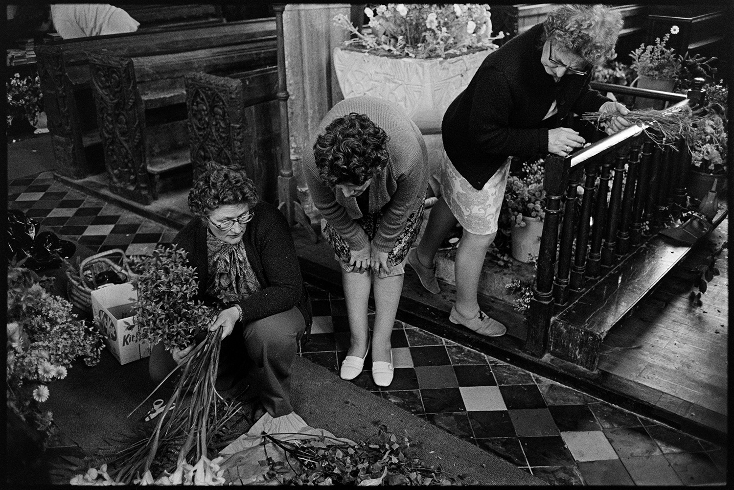 Women decorating church with flowers for Jubilee. Graveyard. 
[Three women decorating High Bickington Church with flowers for Queen Elizabeth II Silver Jubilee. Carved wooden pews can be seen as well as a Norman font with a flower display.]