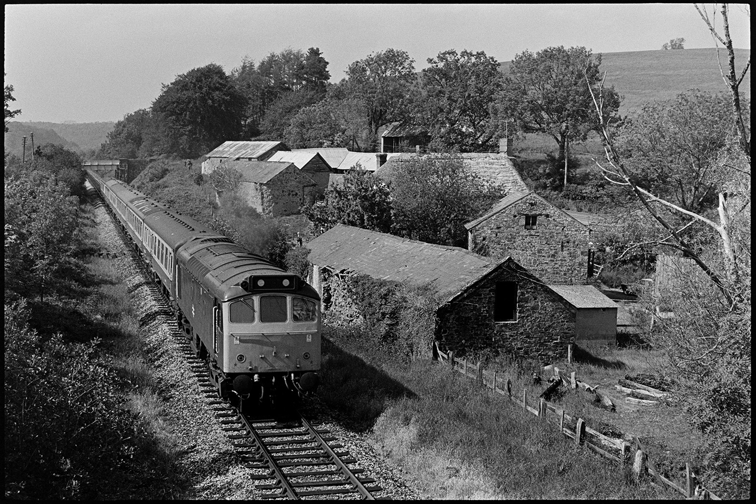 Diesel train on branch line, viewed from above. 
[A diesel train on a branch railway line near Leigh Cross, Chulmleigh. The train is passing a farm.]