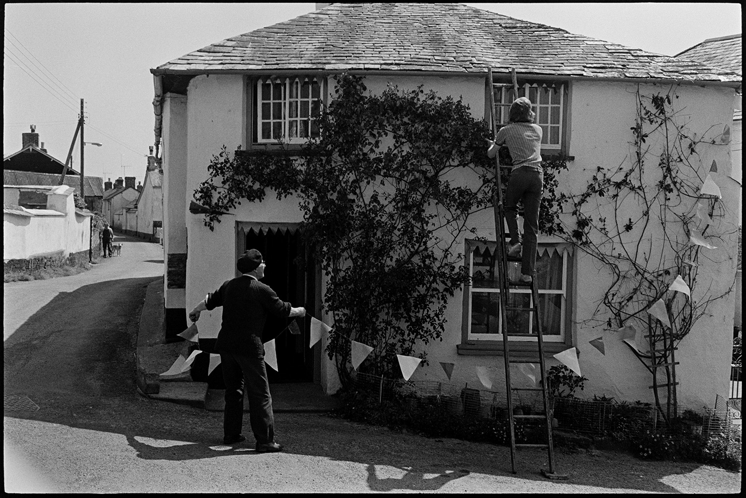 Men putting flags on cottage for Jubilee. 
[Jack Marden, wearing a beret, and another man up a ladder, attaching bunting to Rose Cottage, Aller Road, Dolton for Queen Elizabeth II Silver Jubilee. A rose is growing up the cottage wall.]