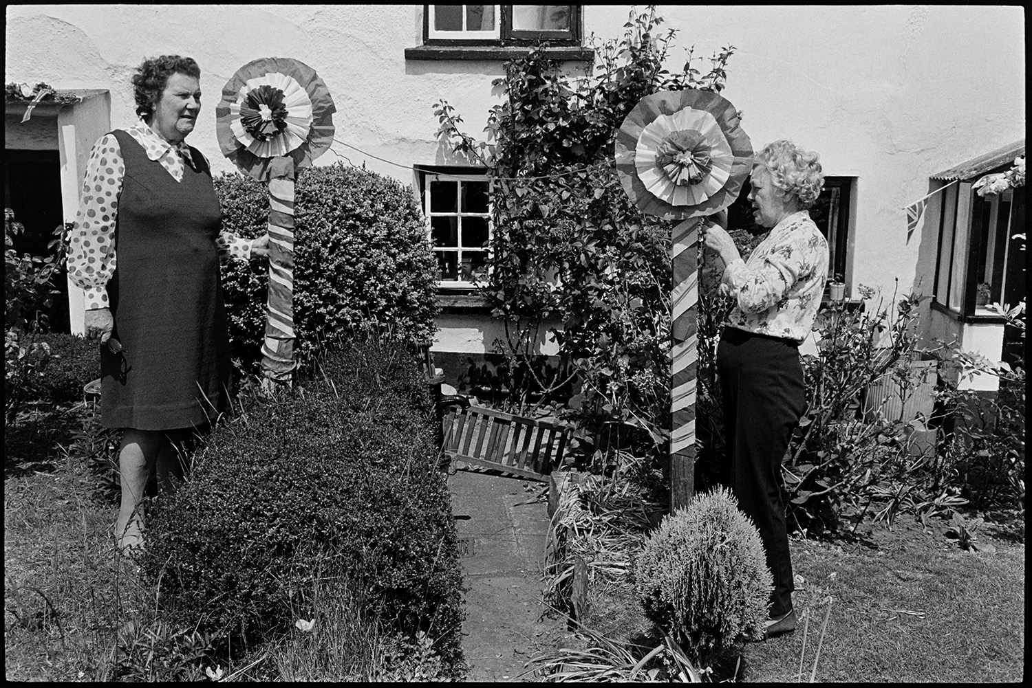 Women standing in garden with decorations for Jubilee. 
[Two women standing in a garden in Dolton with poles they have deorated for Queen Elizabeth II Silver Jubilee. The poles are topped with rosettes.]
