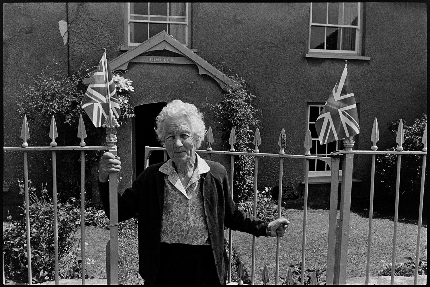 People chatting with flags, decorations for Jubilee. 
[Florence Heaman stood by the gates to Homelea cottage in Dolton. The gates are decorated with Union Jack flags for Queen Elizabeth II Silver Jubilee.]