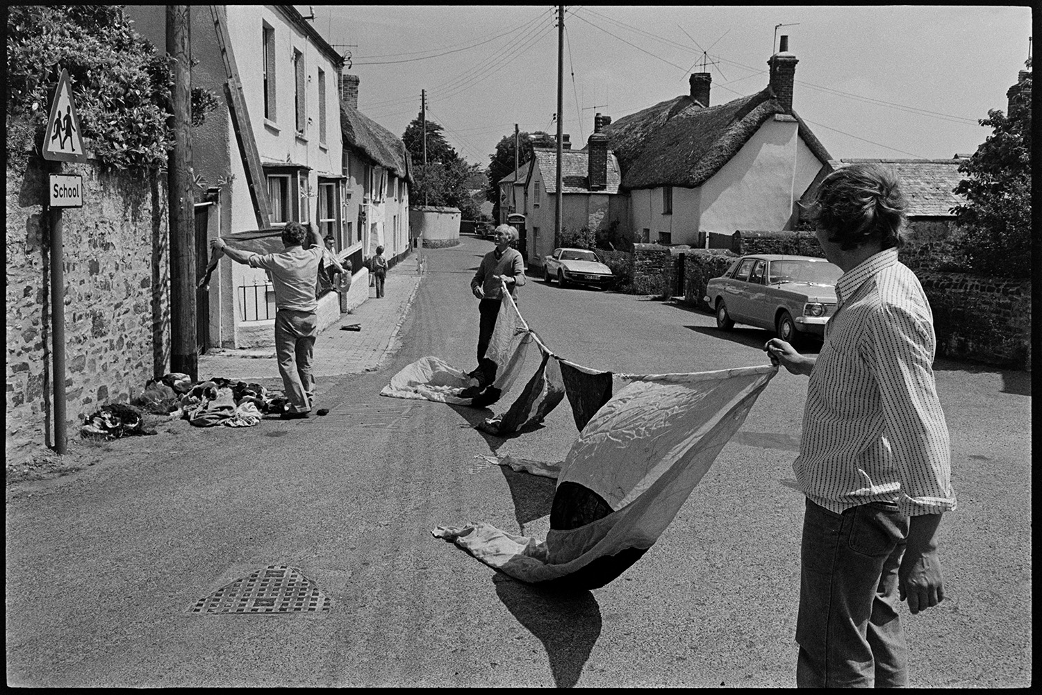 Men putting up large flags, decorations for Jubilee. 
[Fred Folland stood on the right, and two other men putting up flags in Fore Street, Dolton for Queen Elizabeth II Silver Jubilee. A ladder is leant against one of the cottages.]