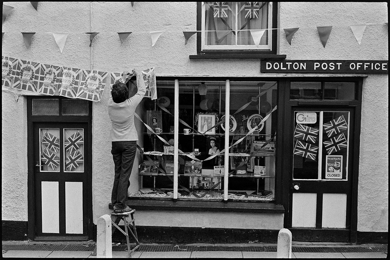 Woman decorating Post Office with flags on Jubilee day! 
[Pat Corbey decorating Dolton Post Office with Union Jack flags, bunting and ribbons on Queen Elizabeth II Silver Jubilee day. The shop window has various royal souvenirs and pictures of the Queen on display.]