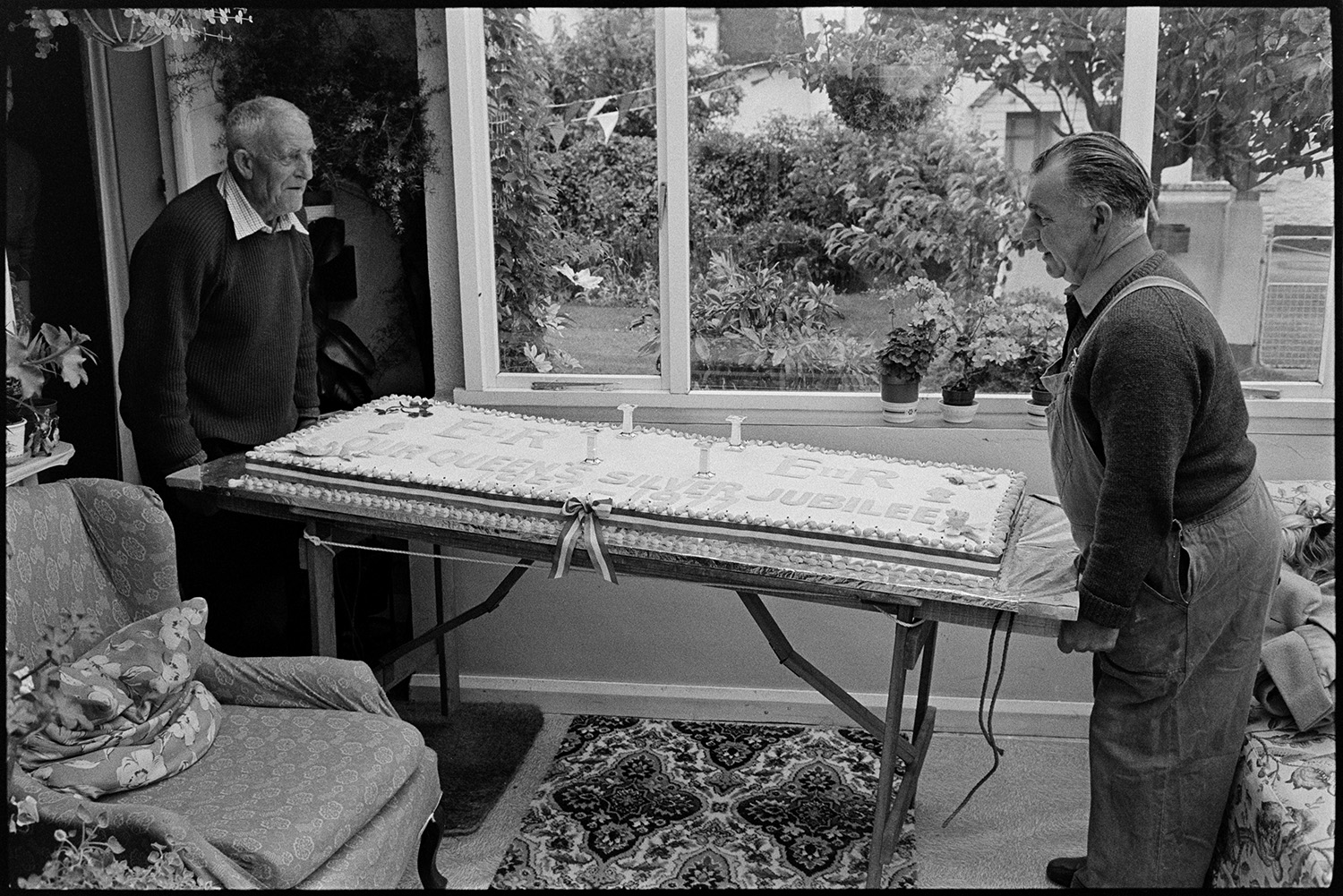 Huge cake being assembled, with crown for tea on Jubilee day! 
[Cyril Dumbleton and another man moving a huge cake through a room in a house in Dolton on Queen Elizabeth II Silver Jubilee Day. The cake is for the village celebrations.]