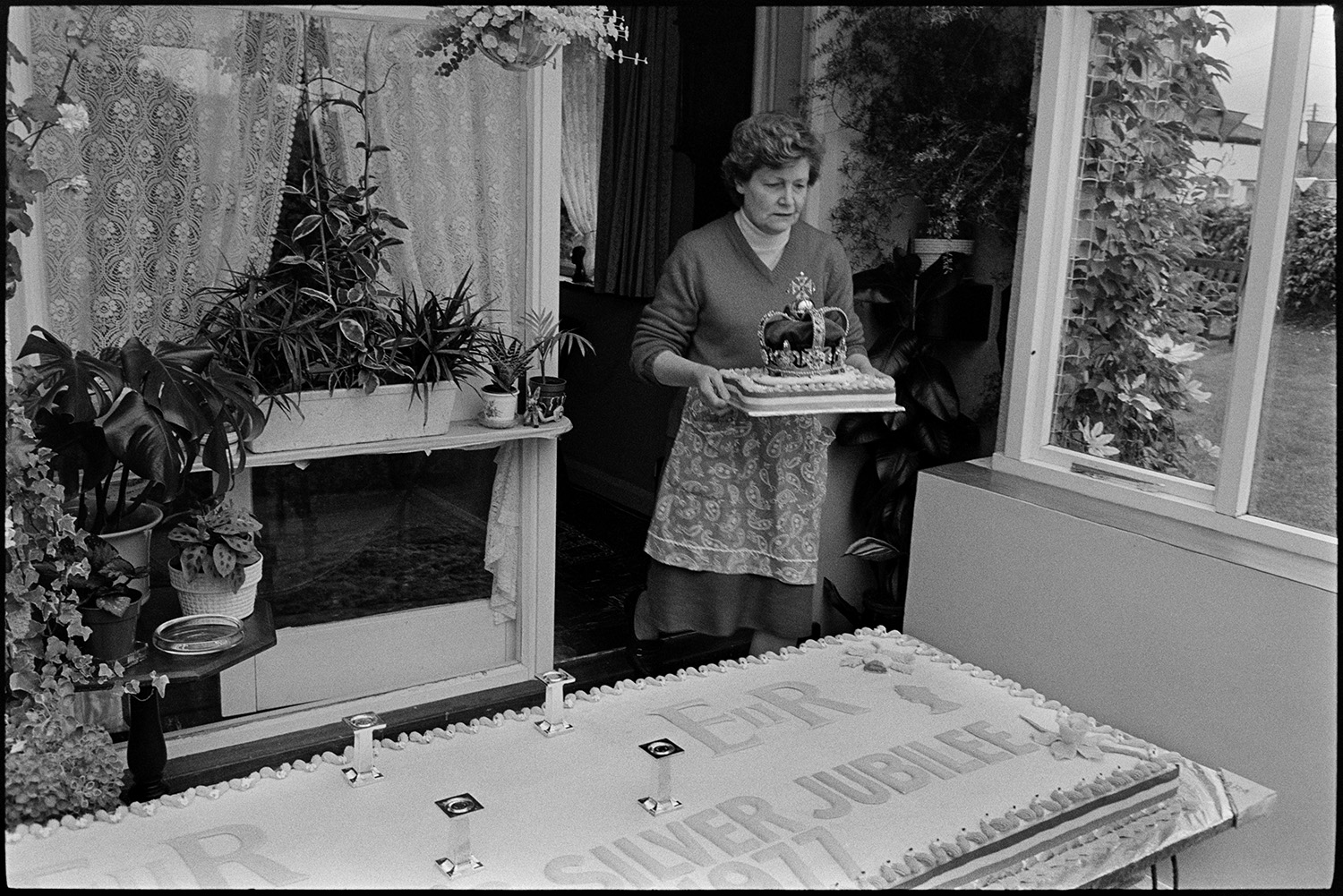 Huge cake being assembled, with crown for tea on Jubilee day! 
[Joan Dumbleton carrying a model crown to place on a huge cake in a house in Dolton to celebrate Queen Elizabeth II Silver Jubilee.]
