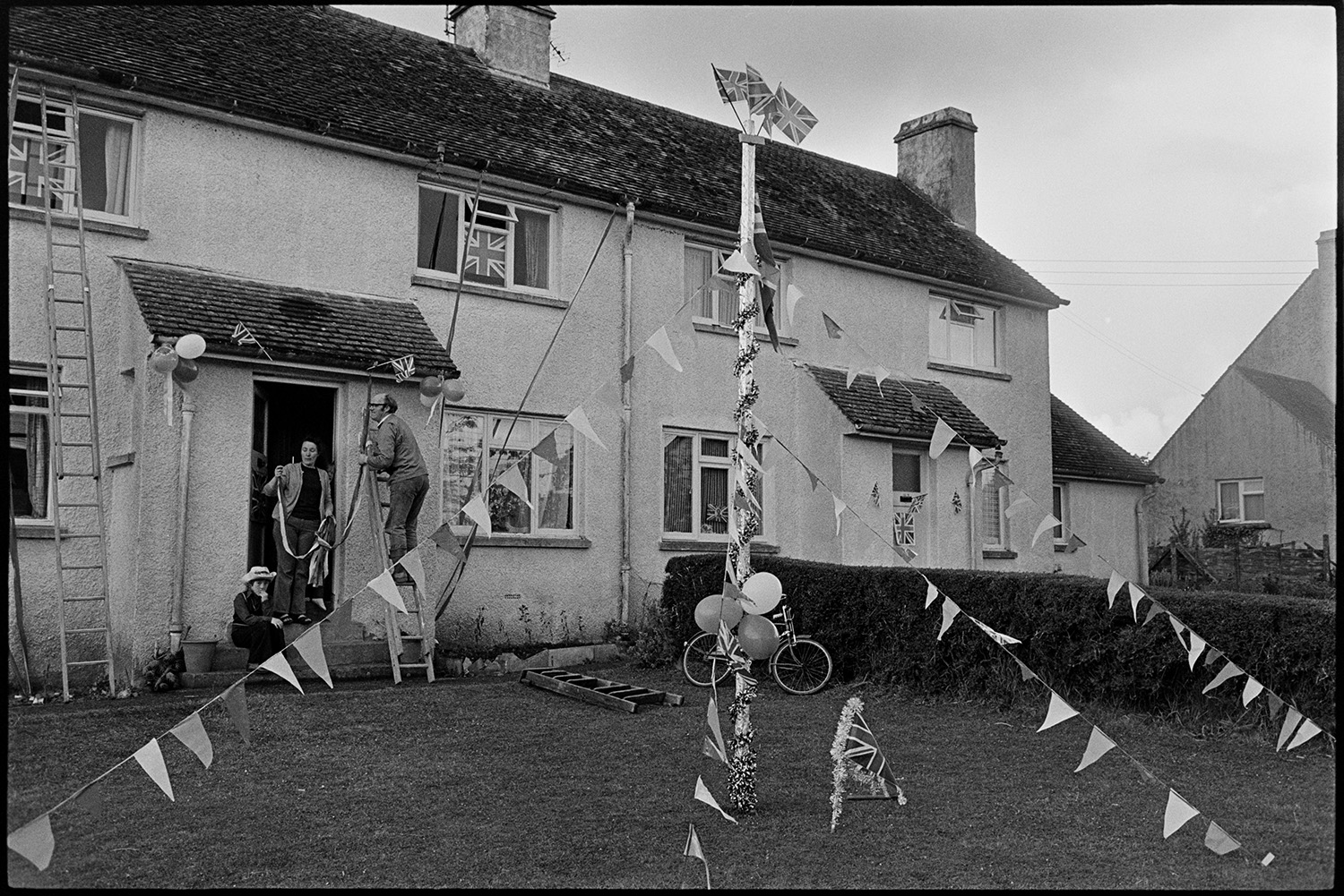 Houses decorated for Silver Jubilee, Electrical shop. 
[A man and woman decorating their house in Dolton with Union Jack flags, balloons and a maypole in the garden on Queen Elizabeth II Silver Jubilee day. Various ladders are propped against the house.]
