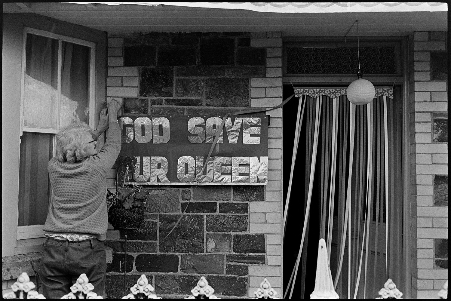 Houses decorated for Silver Jubilee, Electrical shop. 
[A man putting up a banner reading 'God Save Our Queen' on Queen Elizabeth II Silver Jubilee day, in Dolton.]
