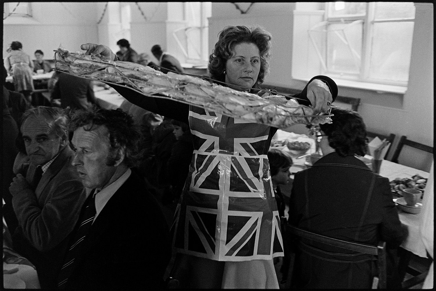 Preparing for Jubilee lunch carrying food. 
[A woman carrying a tray of food at a lunch in Atherington Village Hall to celebrate Queen Elizabeth II Silver Jubilee day.]