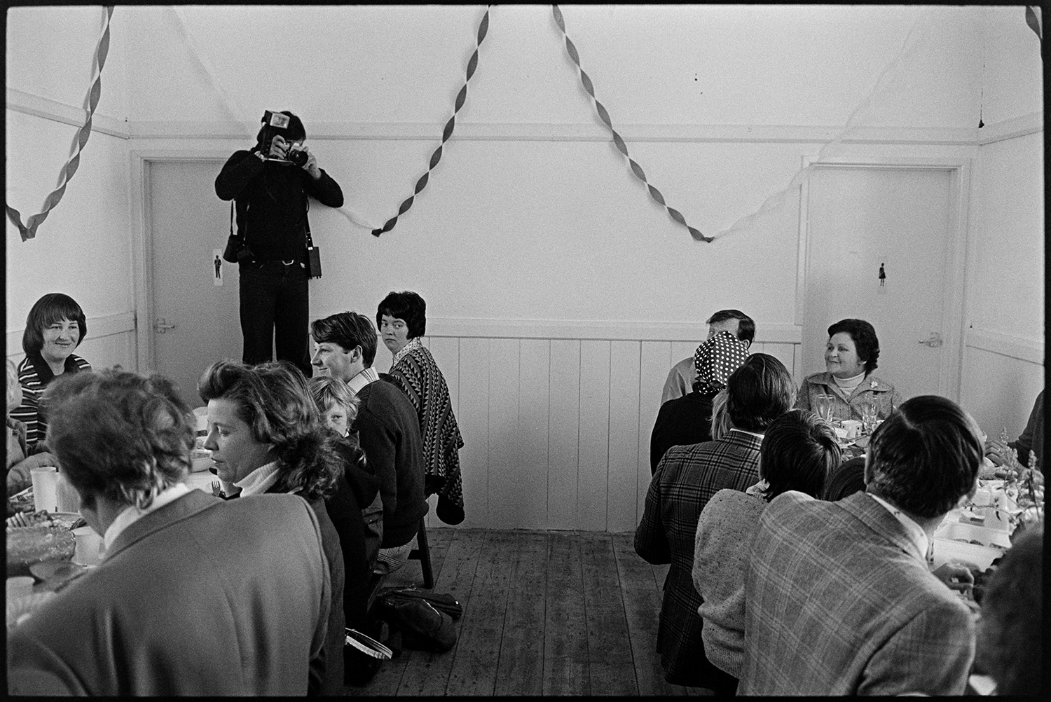 Jubilee lunch in village hall people serving food and eating. 
[A person taking a photograph of people eating lunch in Atherington Village Hall to celebrate Queen Elizabeth II Silver Jubilee day. The hall is decorated with streamers.]