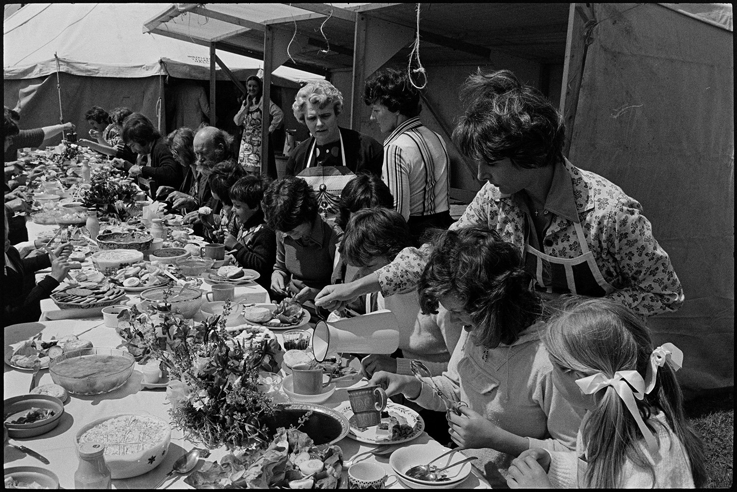 Jubilee lunch in village hall people serving food and eating. 
[Men, women and children eating a lunch to celebrate the Silver Jubilee of Queen Elizabeth II outside Atherington Village Hall. A woman is pouring a drink for one of the children from a jug.]