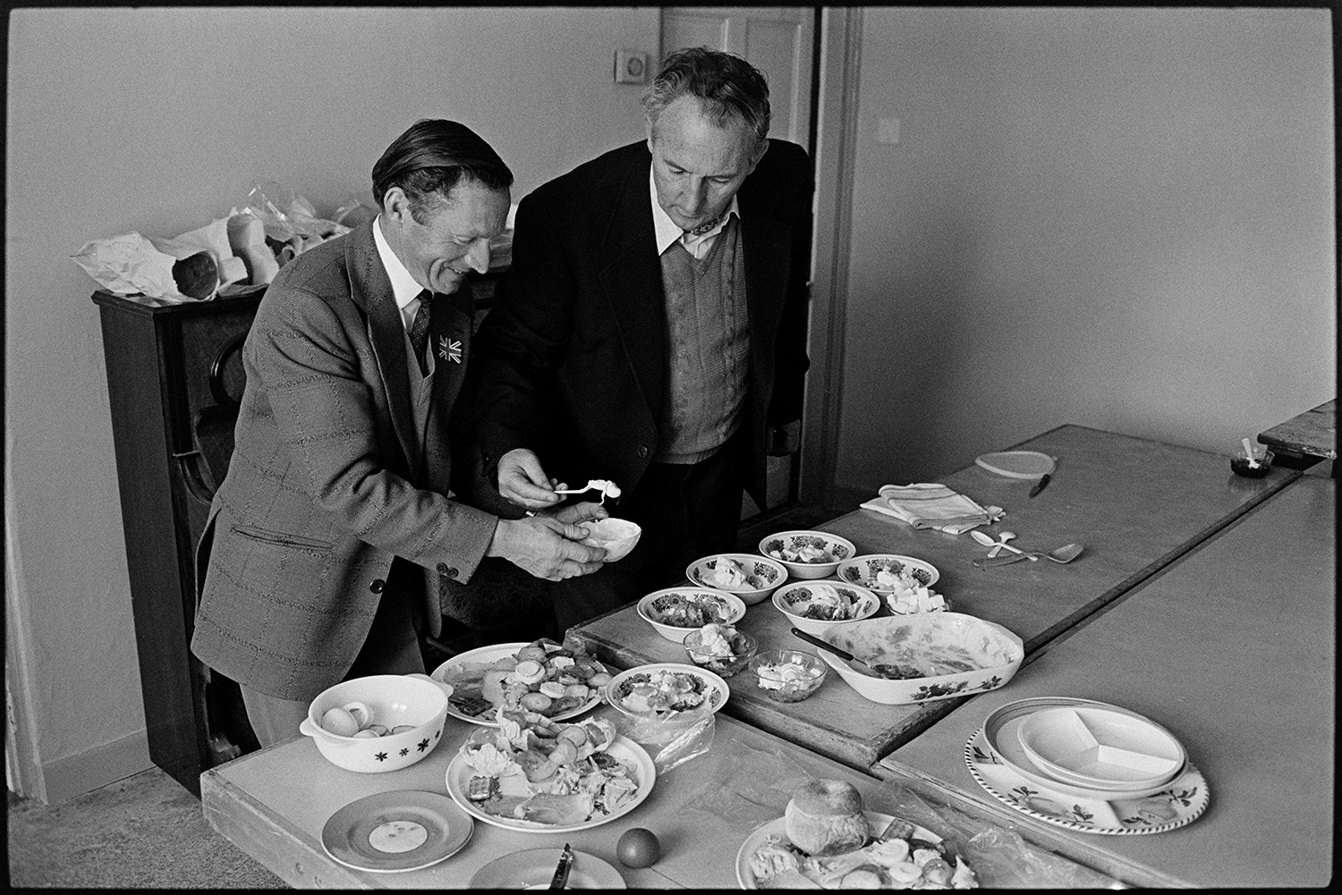 Jubilee lunch in village hall people serving food and eating. 
[Two men adding cream to bowls of desserts on a table in Atherington Village Hall, at a lunch to celebrate the Silver Jubilee of Queen Elizabeth II. Other plates with salads and bread rolls can also be seen on the table.]