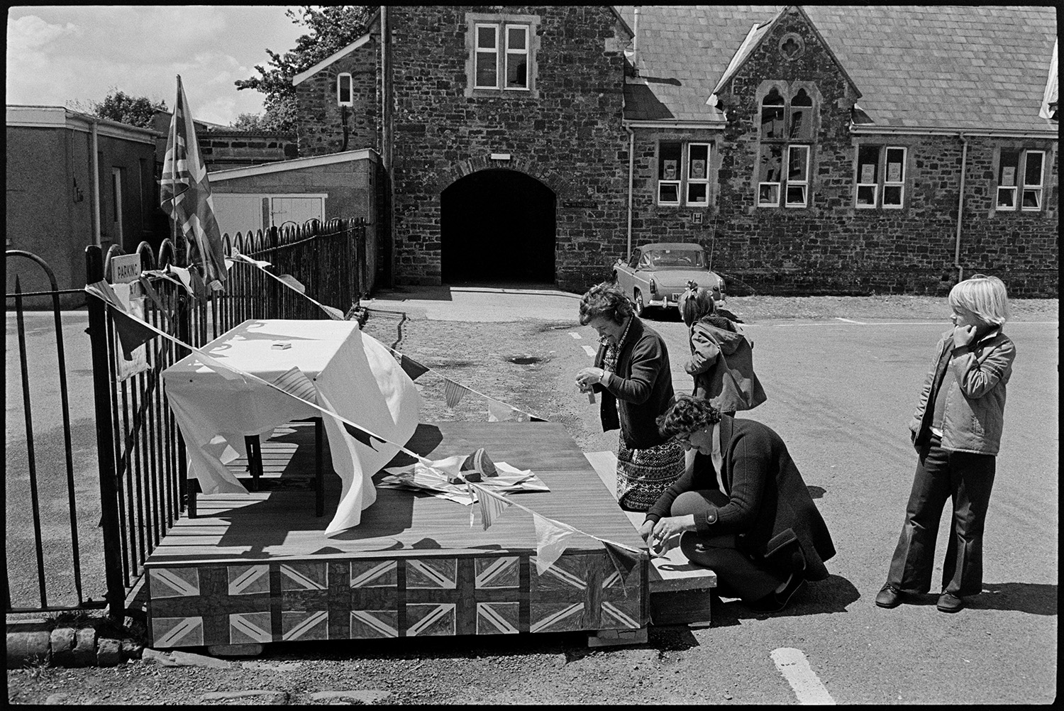 Dais and trestle tables being prepared for Jubilee Day. 
[Two women decorating a small platform and trestle table with bunting and Union Jack flags on the Silver Jubilee of Queen Elizabeth II in High Bickington. Two children are watching them.]