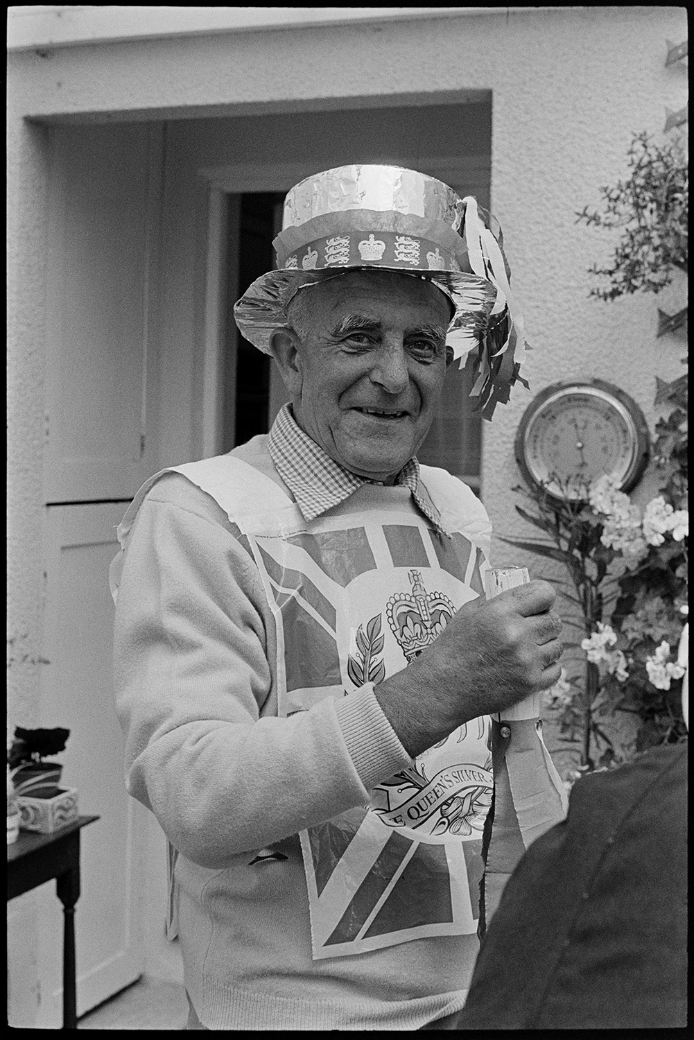 People dressed up for cake procession, Jubilee Day. 
[A man dressed up in a handmade foil  top hat and Union Jack flag for a cake procession in Dolton on the Silver Jubilee of Queen Elizabeth II.]