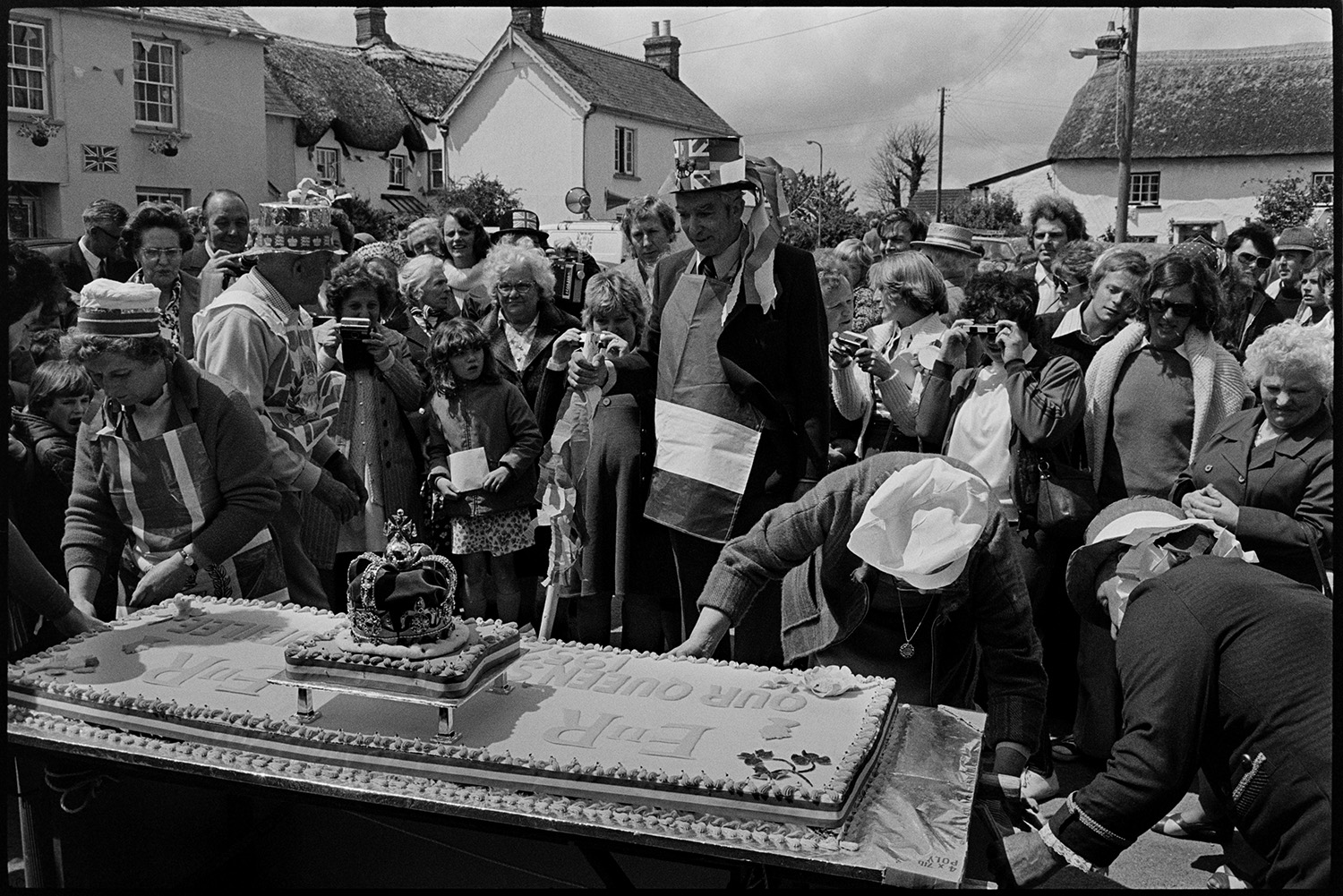 Procession for giant Jubilee cake, village square. Man listening to radio. 
[People securing a giant cake in Dolton village square on the Silver Jubilee of Queen Elizabeth II. A crowd is gathered around the cake and people are taking photographs. Some people are wearing decorated hats and aprons.]