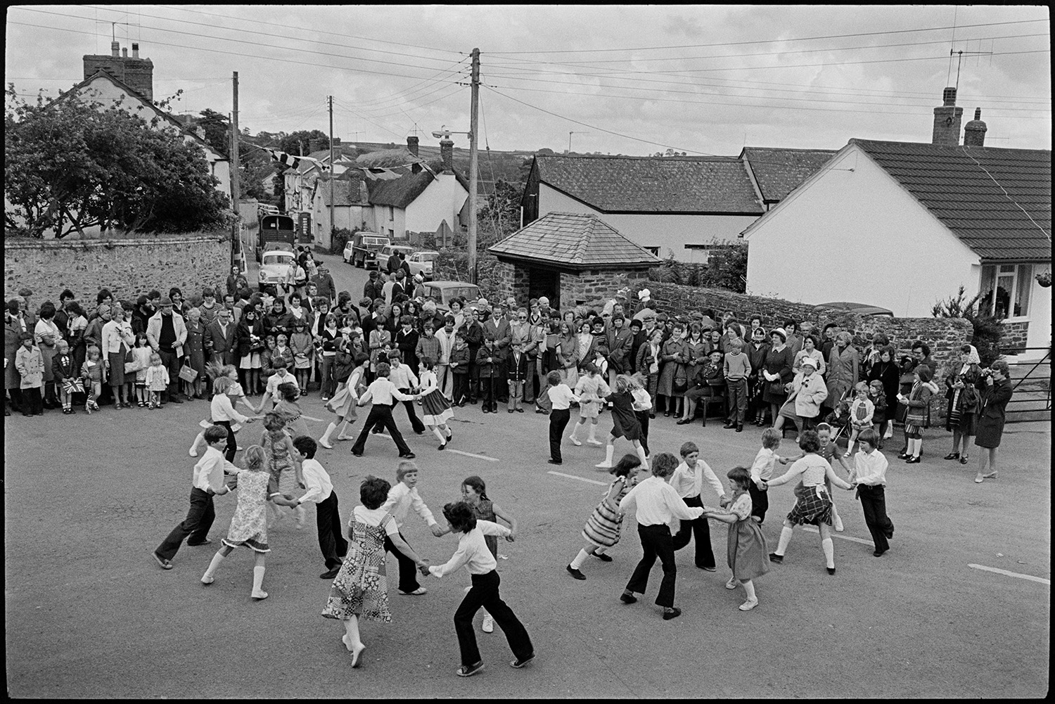 Children dancing in square, Jubilee Day. 
[Children performing a dance in Dolton village square on the Silver Jubilee of Queen Elizabeth II. A crowd of people, stood in front of a bus shelter, are watching them. The street in the background is decorated with flags.]