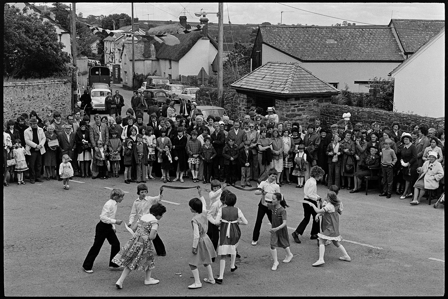 Children dancing in square, Jubilee Day. 
[Children performing a dance with ribbons in Dolton village square on the Silver Jubilee of Queen Elizabeth II. A crowd of people, stood in front of a bus shelter, are watching them. The street in the background is decorated with flags.]