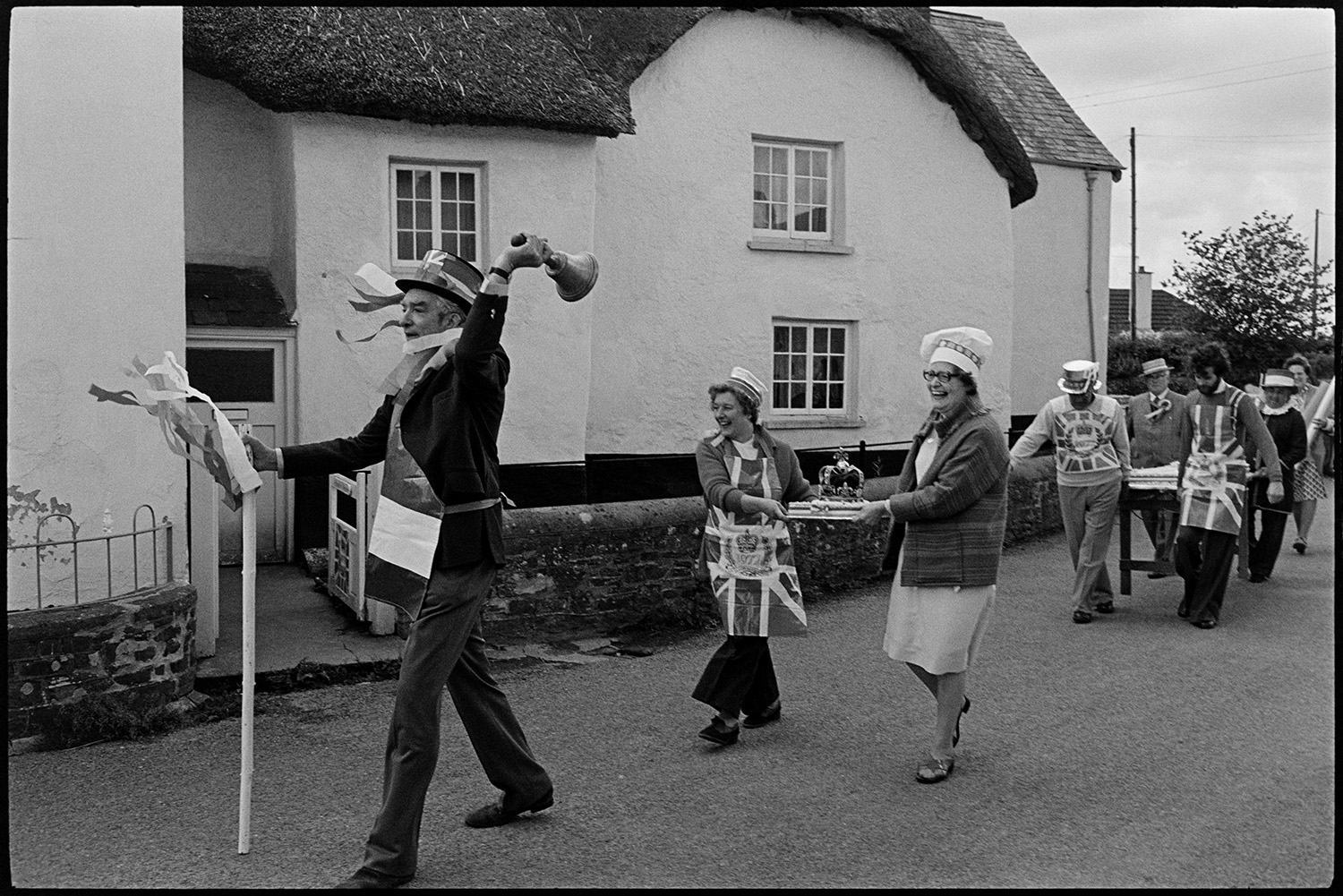 Procession for giant Jubilee cake, village square. Man listening to radio. 
[Men and women parading a giant cake through a street in Dolton to celebrate the Silver Jubilee of Queen Elizabeth II. A man leading the procession, possibly a town crier, is ringing a bell. They are passing a thatched cottage and are wearing aprons with Union Jack designs.]