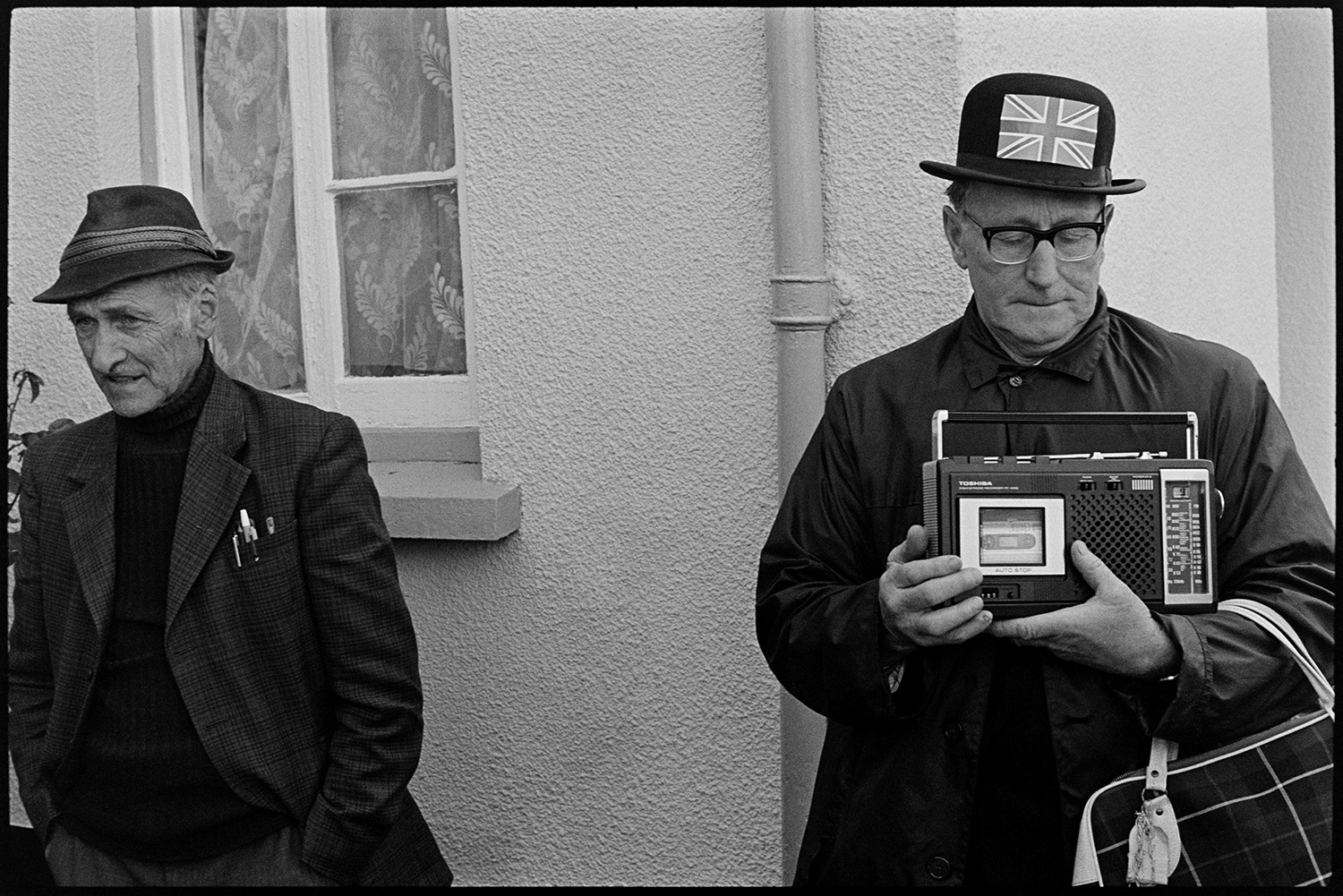 Spectators in square, Jubilee Day, man with radio. 
[Jack Marden holding a tape recorder and watching the celebrations for the Silver Jubilee of Queen Elizabeth II in Dolton village square. He is wearing a hat decorated with a Union Jack flag and stood next to another man.]