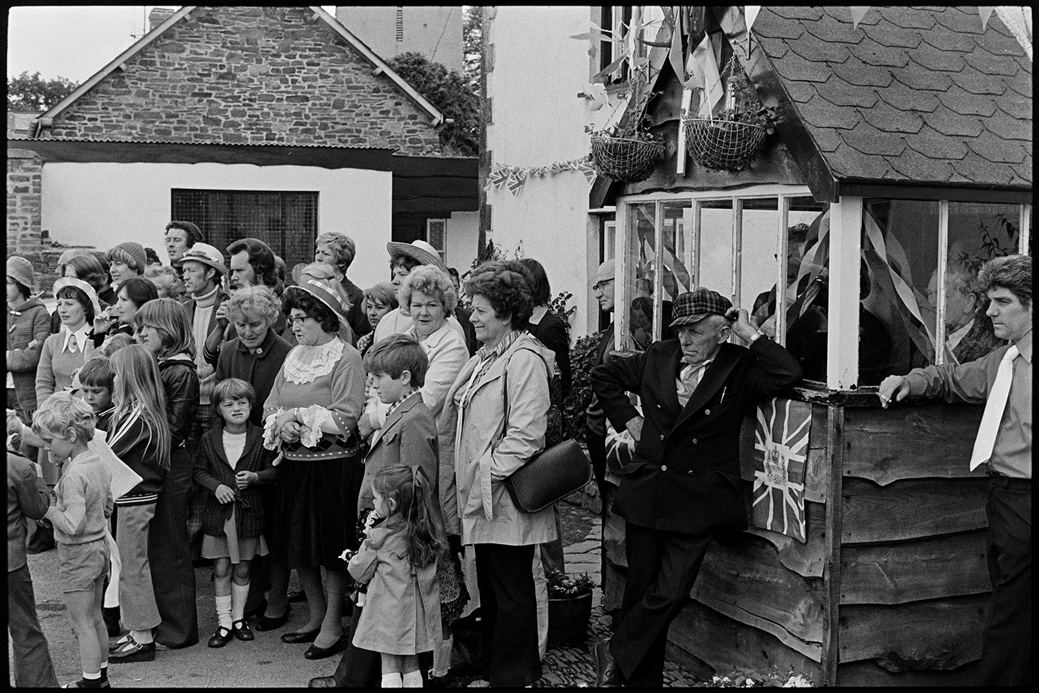 Spectators in square, Jubilee Day, man with radio. 
[Men, women and children gathered outside the Royal Oak pub in Dolton to watch the celebrations for the Silver Jubilee of Queen Elizabeth II in the village square. The pub is decorated with Union Jack flags and bunting.]