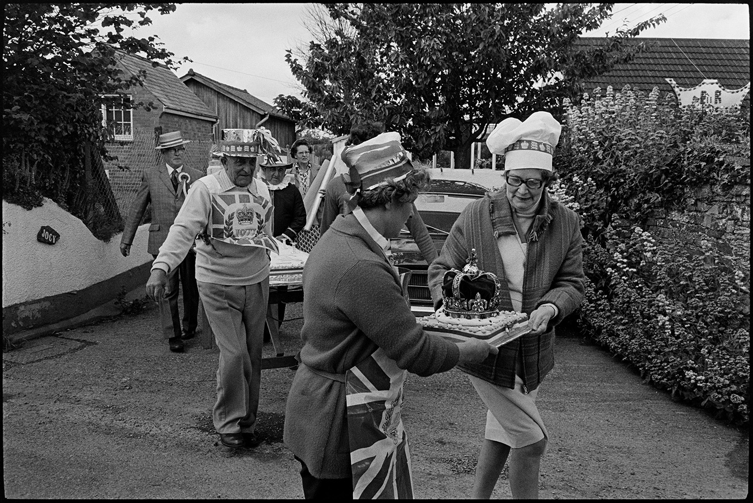 Procession for giant Jubilee cake, village square. Man listening to radio. <br />
[Men and women parading a giant cake through a street in Dolton to celebrate the Silver Jubilee of Queen Elizabeth II. The people are wearing decorated hats and aprons.]