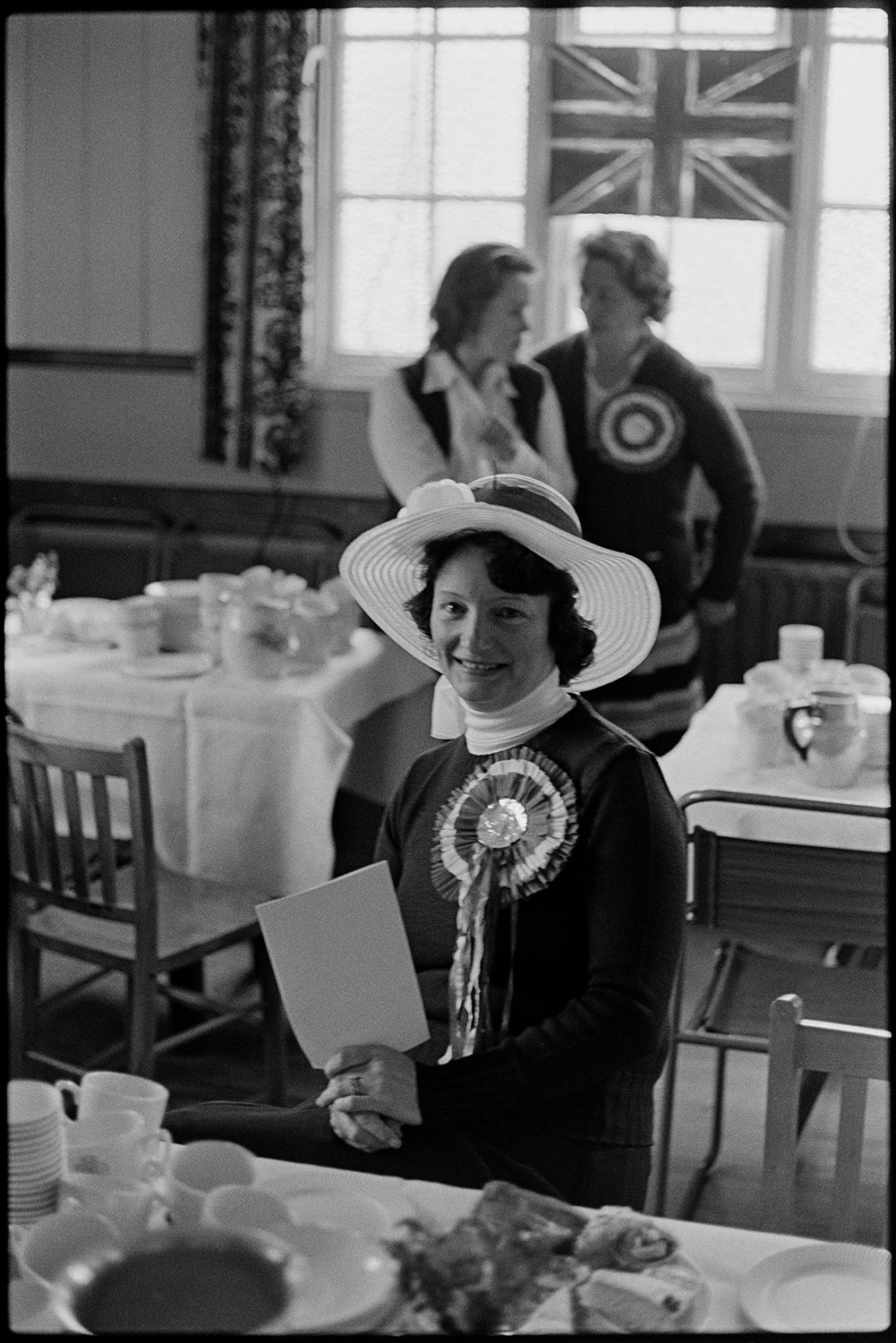Group of women, in village hall before Jubilee tea which they had prepared. 
[A woman sat at a table laid with tea and cake, which she helped to prepare, in High Bickington Village Hall to celebrate the Silver Jubilee of Queen Elizabeth II.  She is wearing a hat and rosette. Two other women are stood in the background by a Union Jack flag.]