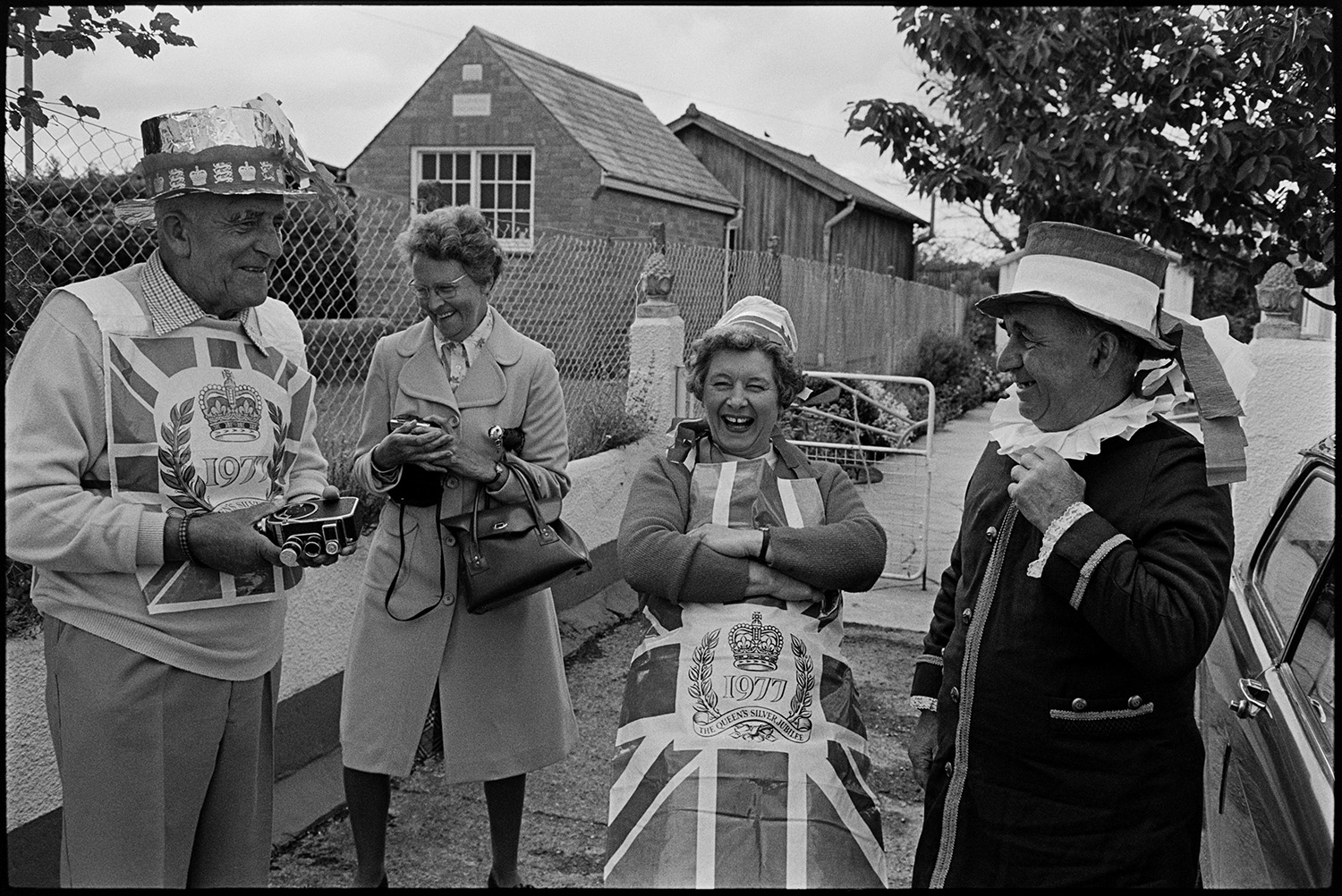 People chatting after Jubilee cake ceremony. 
[Two men and two women talking and laughing after parading a giant cake through Dolton to celebrate the Silver Jubilee of Queen Elizabeth II. Dorothy Hiscock I stood in the centre wearing an apron with a Union Jack motif. The men are wearing decorated hats and one of them is holding a camera.]