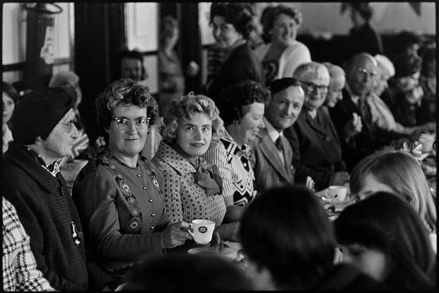 Jubilee tea in village hall photographer at work. 
[Women and men sat at a table in High Bickington Village Hall, at a tea to celebrate the Silver Jubilee of Queen Elizabeth II. Mr and Mrs Thomas are sat in the centre of the picture, fourth and fifth from the front.]