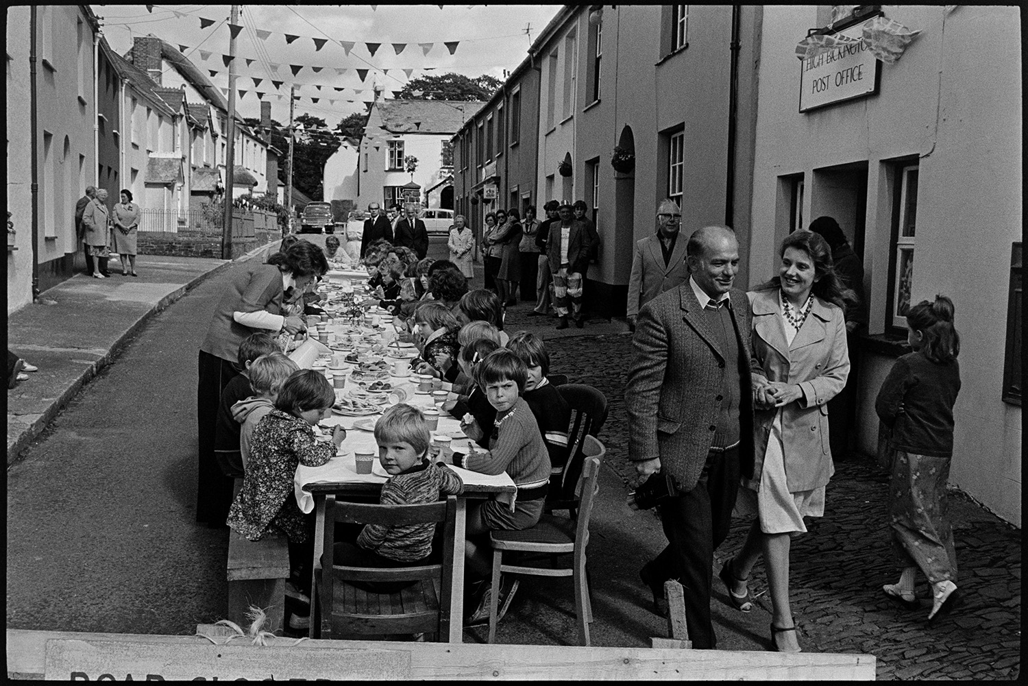 Children's tea in village street, Jubilee Day. 
[Children sat a trestle tables having tea in the street outside High Bickington Post Office to celebrate the Silver Jubilee of Queen Elizabeth II. The street is decorated with bunting and people are walking past.]