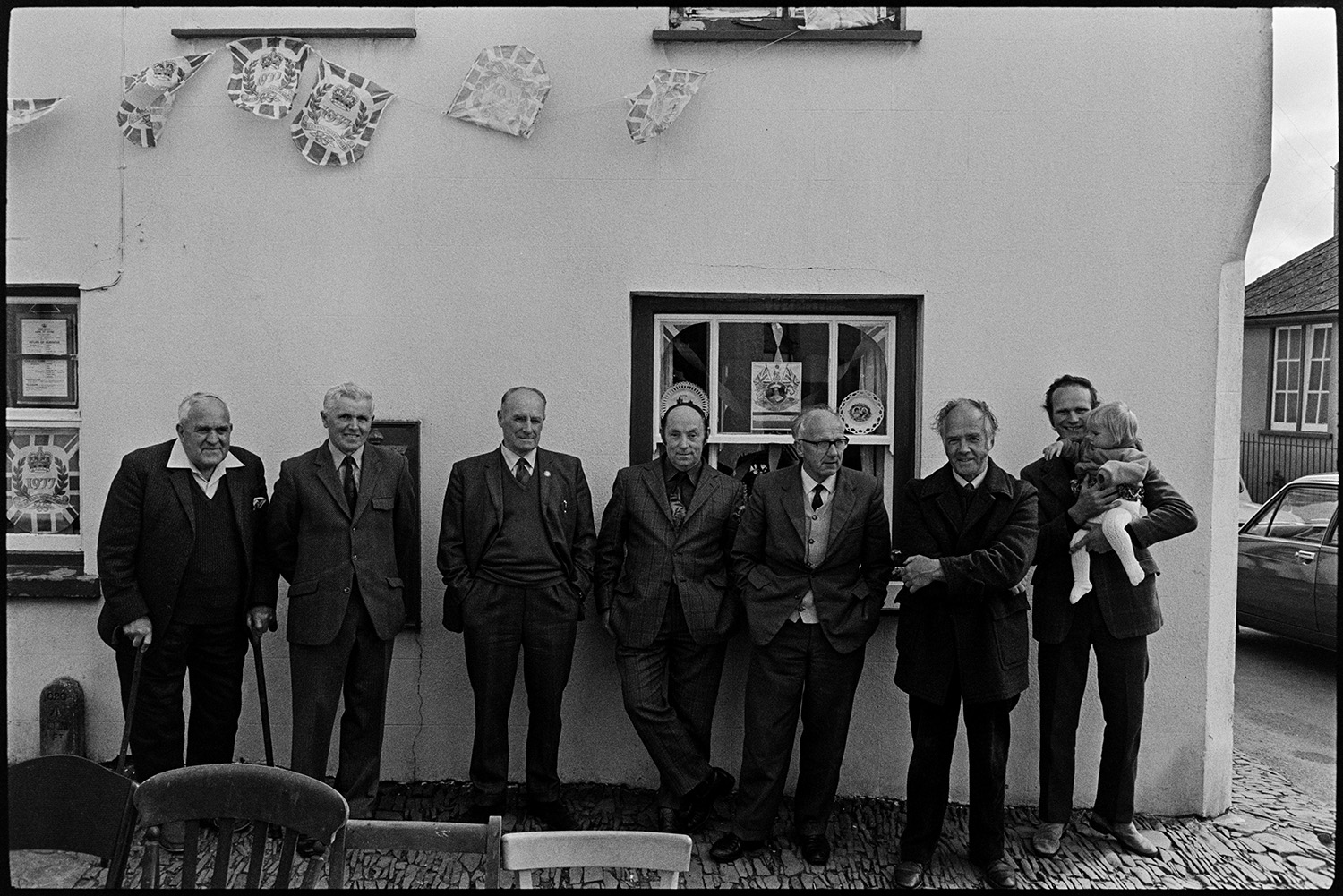 Men waiting for start of Jubilee tea in village street. 
[A row of men stood in a street in High Bickington waiting for the start of a tea or street party to celebrate the Silver Jubilee of Queen Elizabeth II. One of the men is holding a child and the building behind is decorated with bunting. John Tucker is second from left and Bill Laramy is third from the left. Mr Marden is third from the right.]