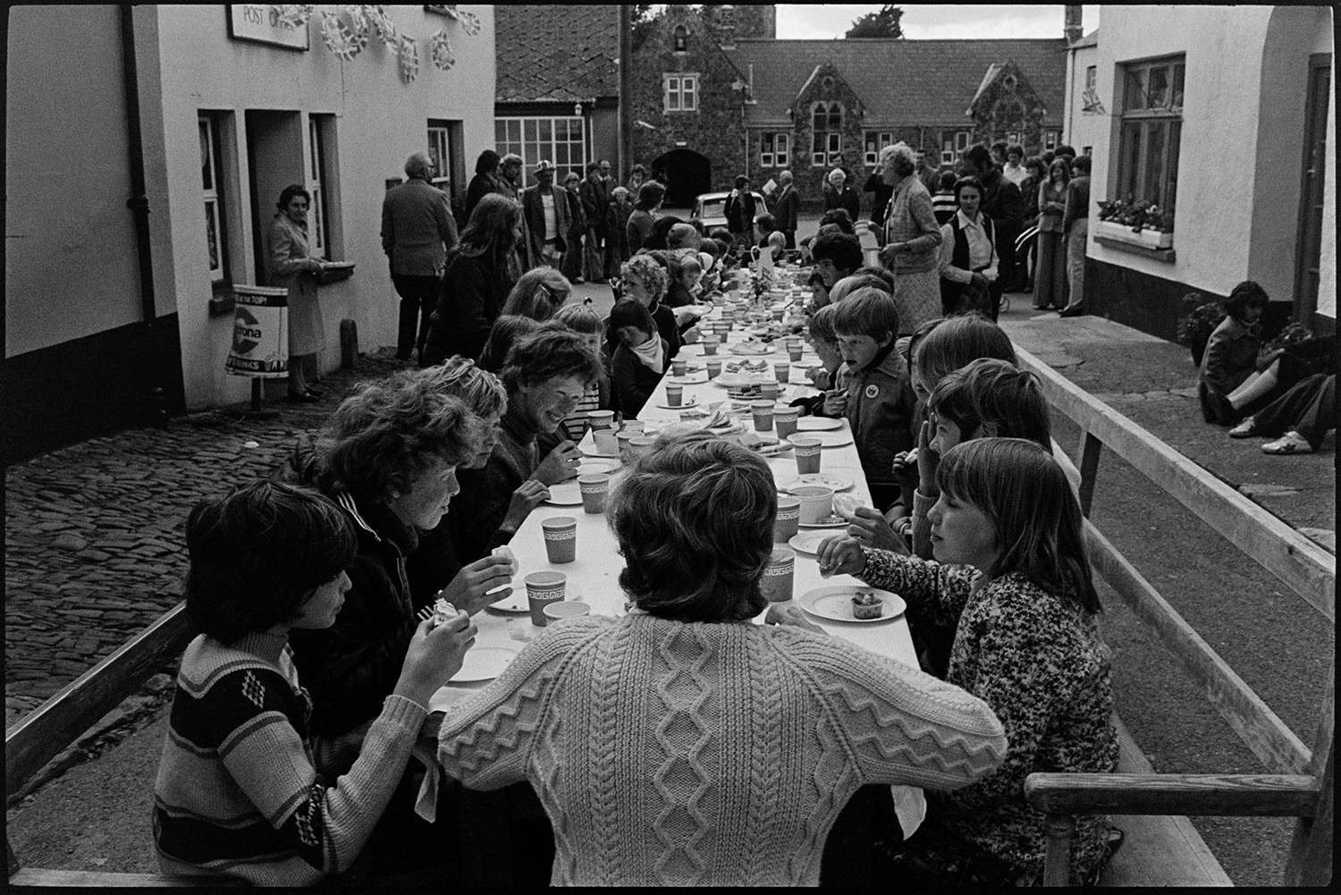 Children's tea in village street, Jubilee Day. 
[Children sat a trestle tables having tea in the street outside High Bickington Post Office to celebrate the Silver Jubilee of Queen Elizabeth II. The Post Office is decorated with bunting and people are stood in the street. High Bickington Primary School can be seen in the background.]