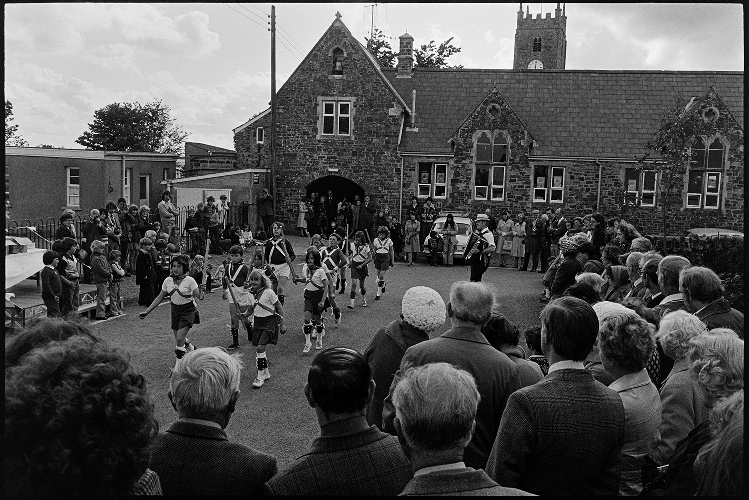 Children Morris dancing in village street. 
[Child Morris Dancers performing in front of a crowd outside High Bickington Primary School as part of the celebrations for the Silver Jubilee of Queen Elizabeth II. A man is playing the accordion for them.]