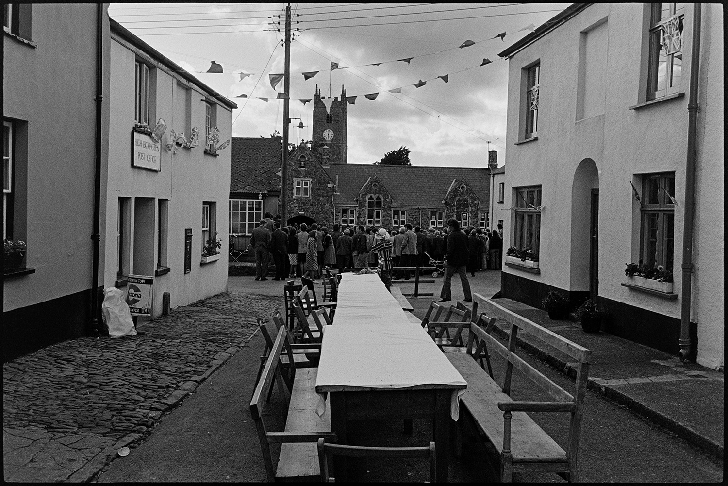 Empty tables in street after Jubilee tea, crowd watching dancing in background. 
[Empty trestle tables and chairs outside High Bickington Post Office after a tea to celebrate the Silver Jubilee of Queen Elizabeth II. The street is decorated with bunting. In the background a crowd is watching child Morris Dancers perform in front of High Bickington Primary School. High Bickington church tower and clock can be seen in the distance.]