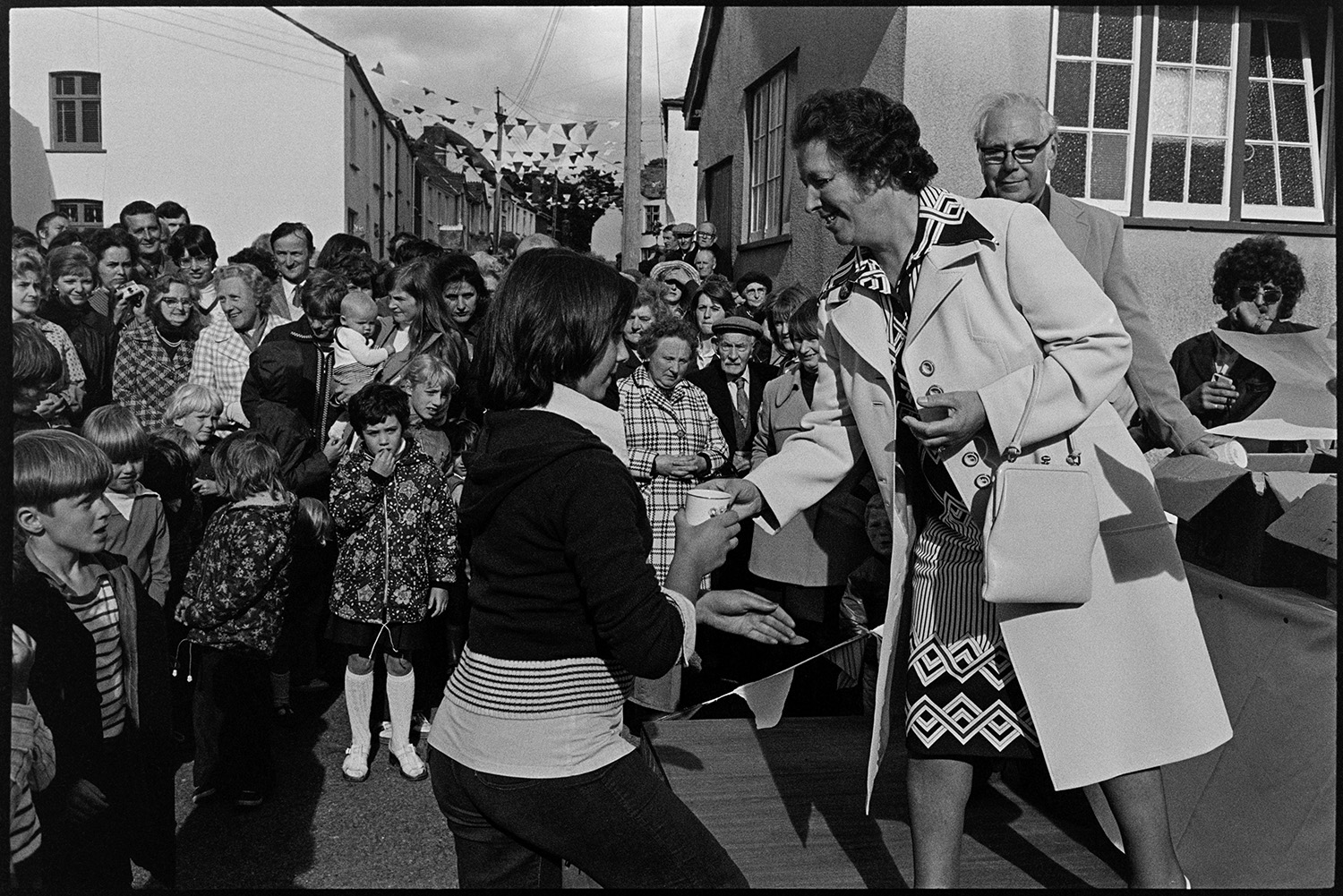 Presentation of mugs in street, crowd watching. 
[Mrs Thomas presenting mugs to children in a street in High Bickington to celebrate the Silver Jubilee of Queen Elizabeth II. A crowd is watching the presentation. The street in the background is decorated with bunting.]